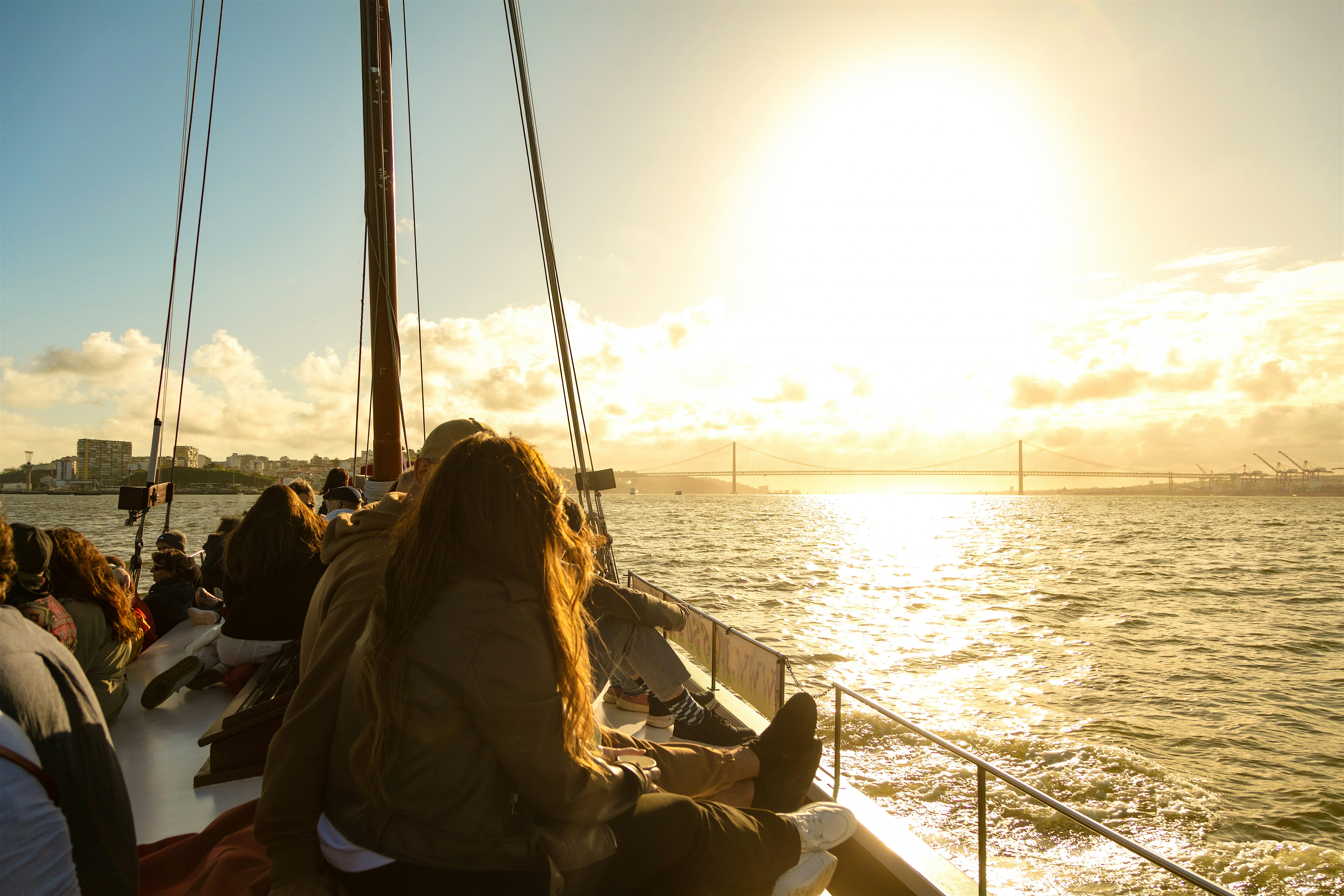 People seated along the side of a boat watching the sun set over shimmering water, with a long suspension bridge stretching across the horizon.