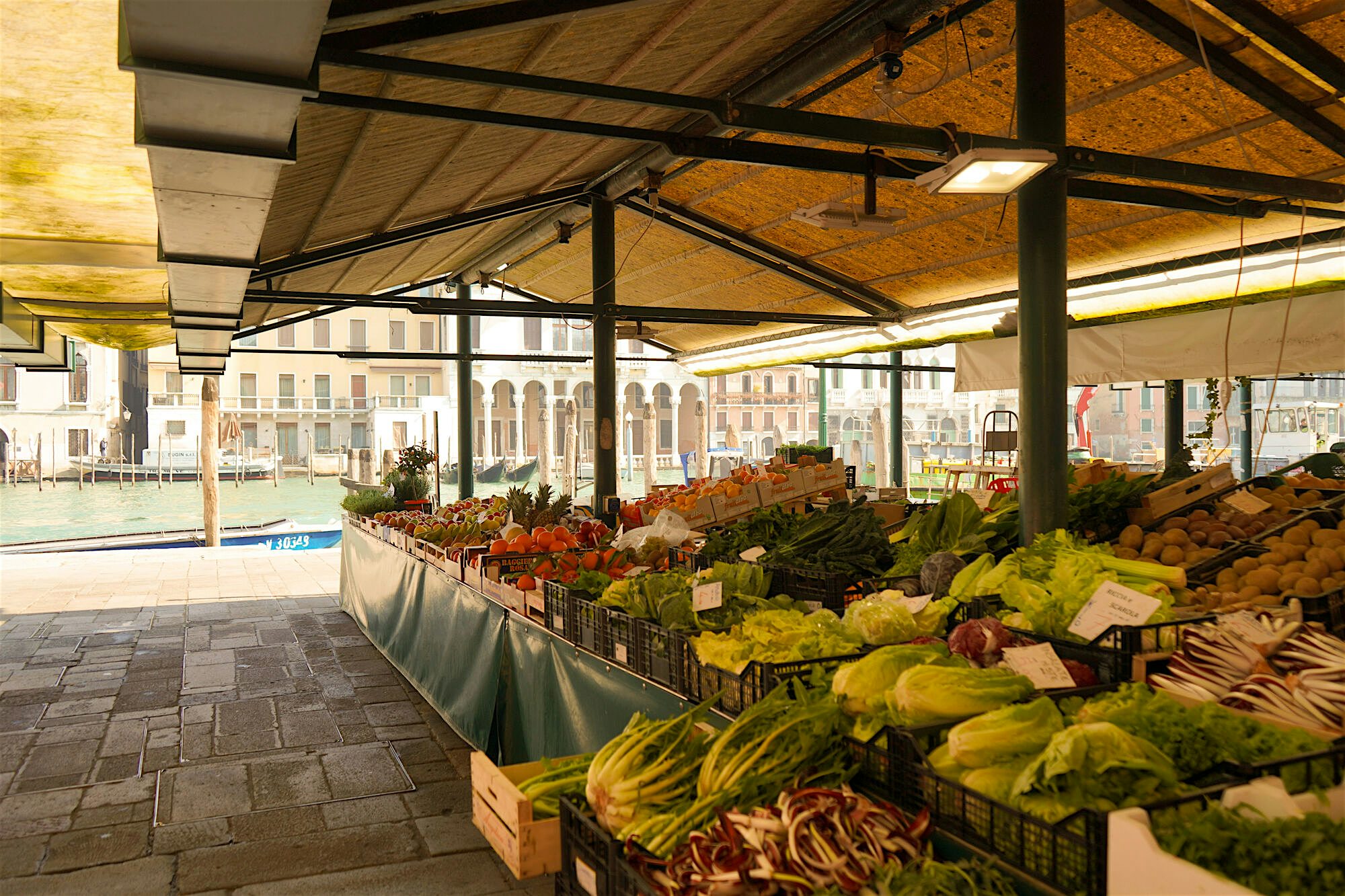 "A covered market stall displaying fresh vegetables and produce beside a canal, with buildings reflected in the water.  "