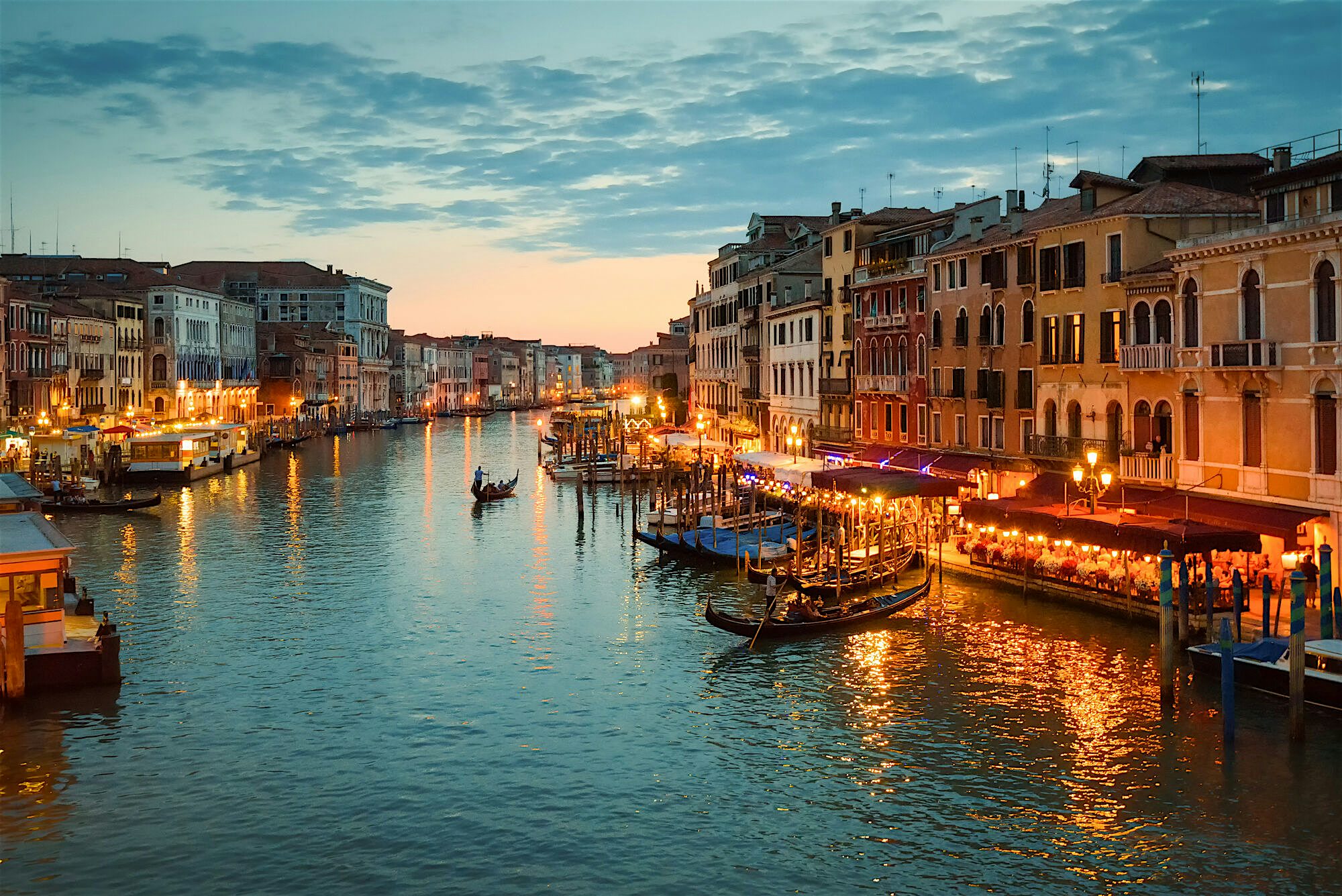 A birds eye view of a Venitian canal at sunset. Bars, and restauants along the waterside light up with a range of lamps and warm lighting. One Gondola can be seen travelling.