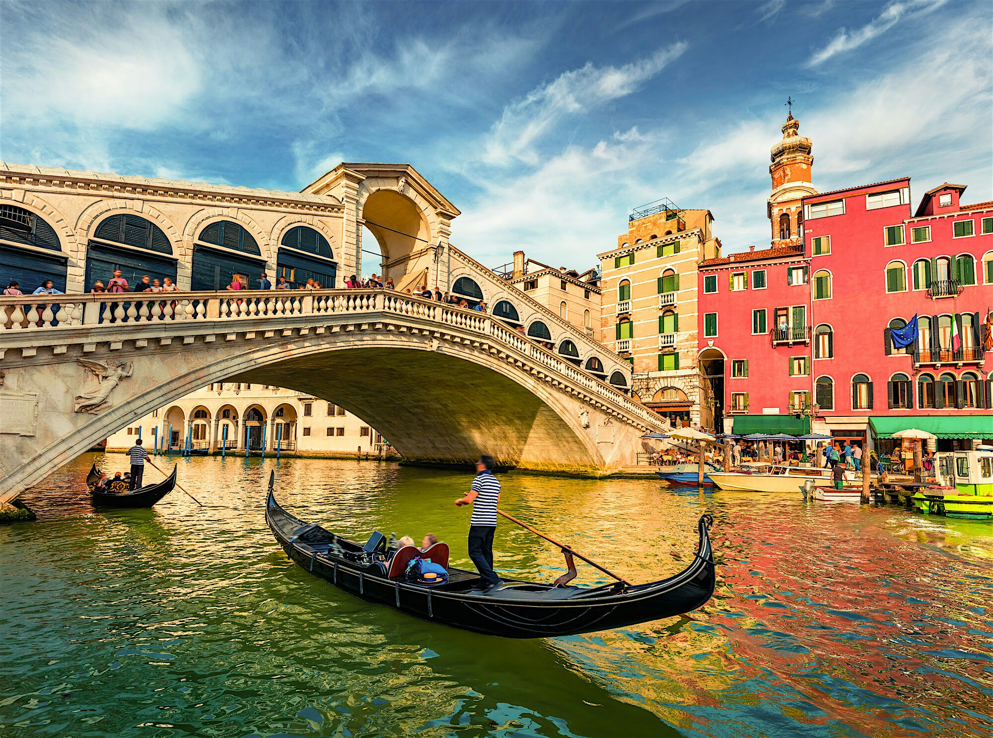 A canal with a number of gondolas riding under the Rialto Bridge, the oldest bridge on Venices canal network. Tall bright buildings line the street beside and crowds upon the bridge and pathway.