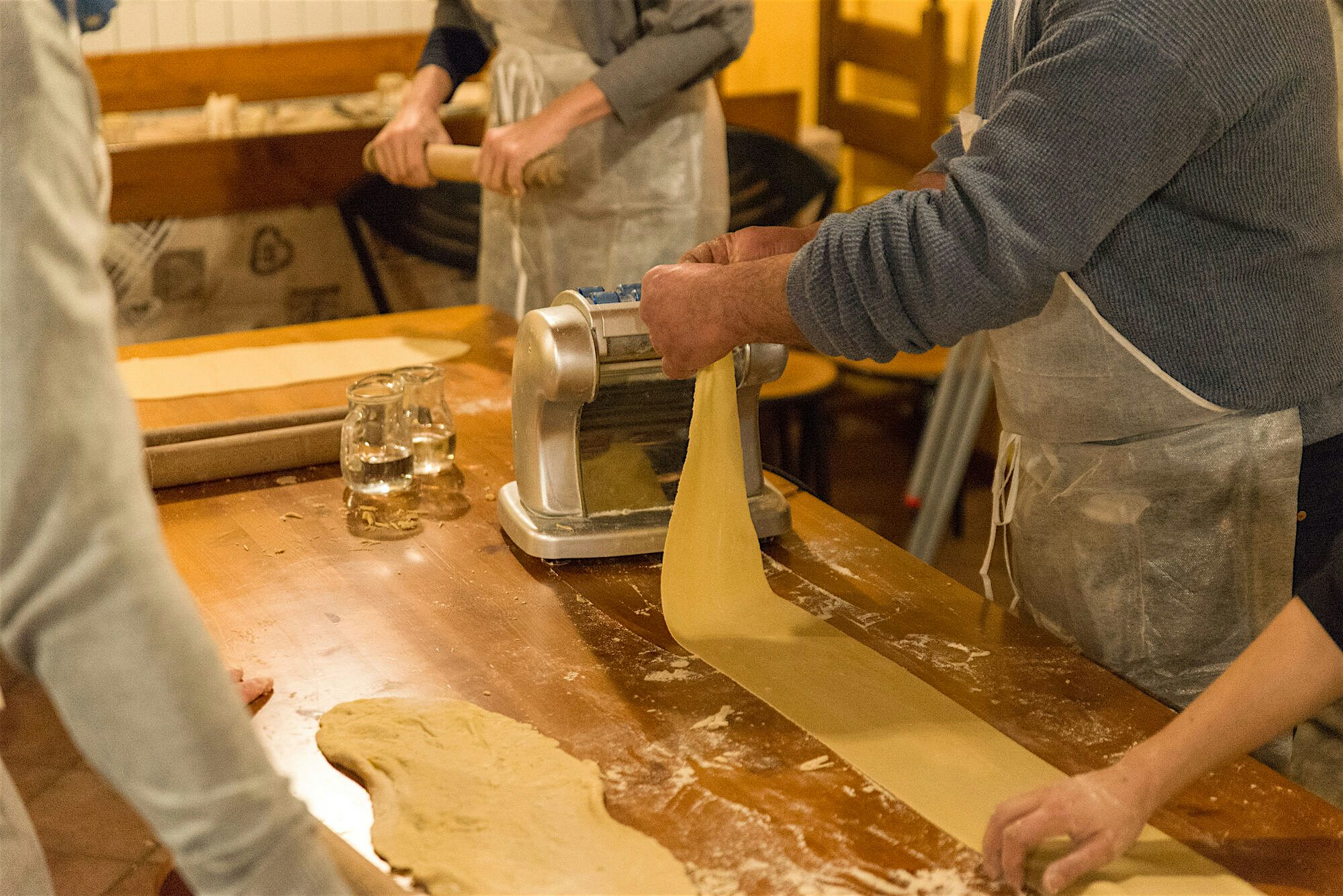 A pasta making workshop, one chef in a grey apron loads up a dough into a roller, and other stand watching with a hand roller in hand