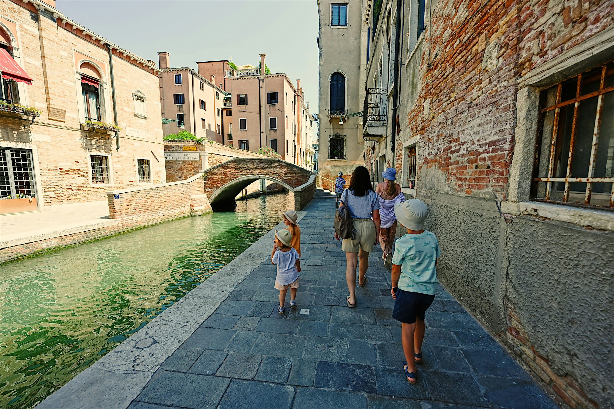 A family walks down the path on an old street by the canal. The sky is bright, and clear behind them