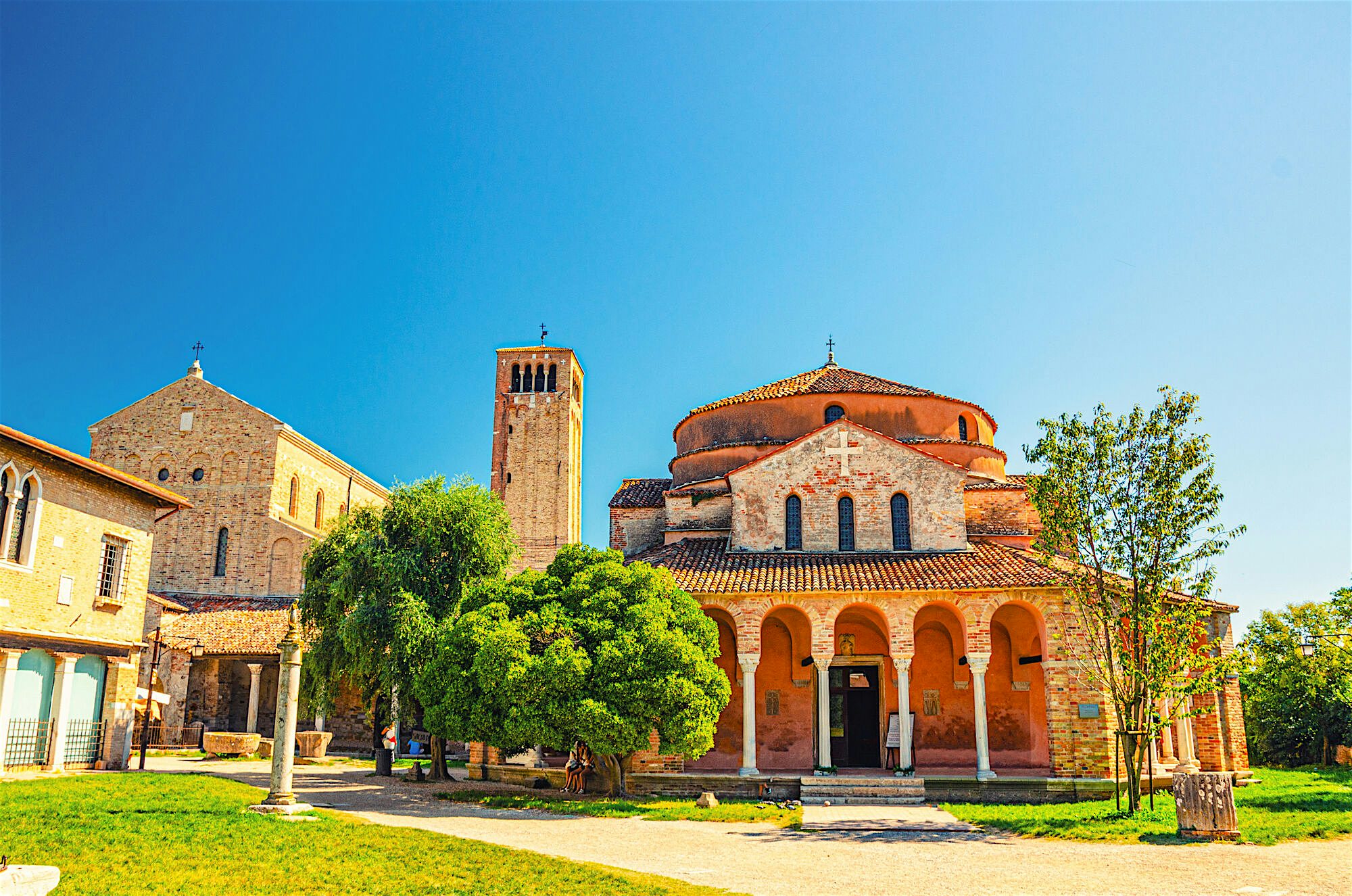"A historic brick church with arched entrances set within a green courtyard, surrounded by trees under a clear blue sky.  "