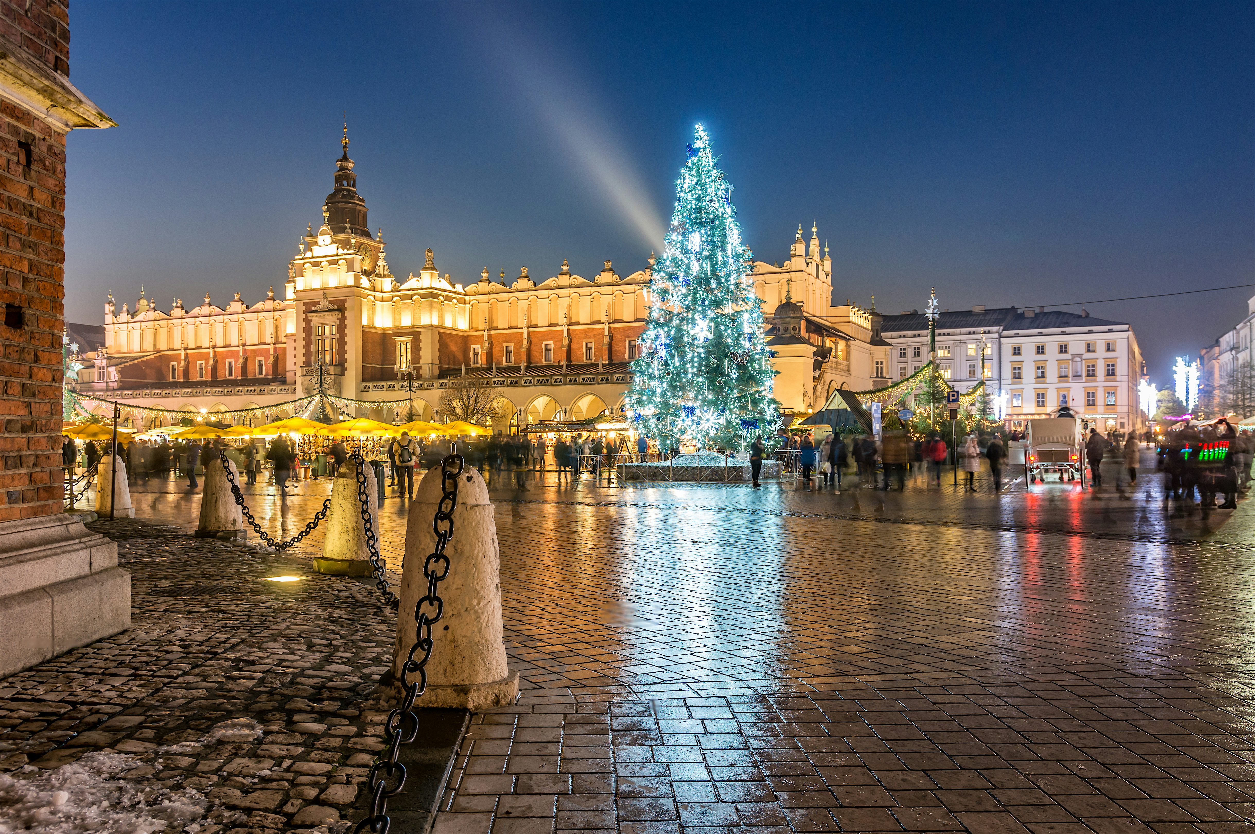 Illuminated city square at night with a large decorated Christmas tree at the centre, surrounded by market stalls and people, and backed by an ornate historic building glowing