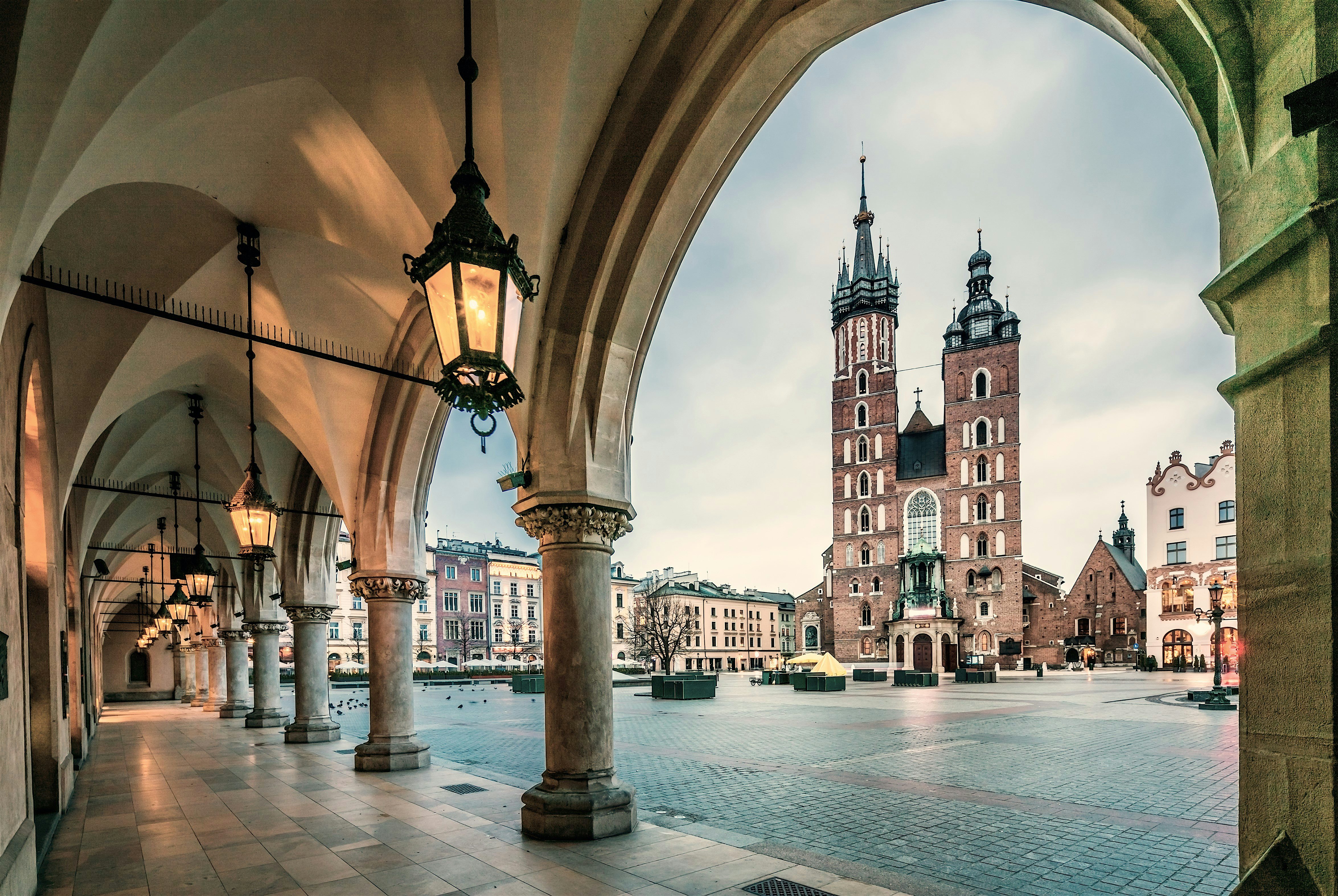 View through a covered stone arcade with vaulted ceilings and hanging lanterns, opening onto a wide historic square dominated by a twin‑towered brick church and surrounding city buildings