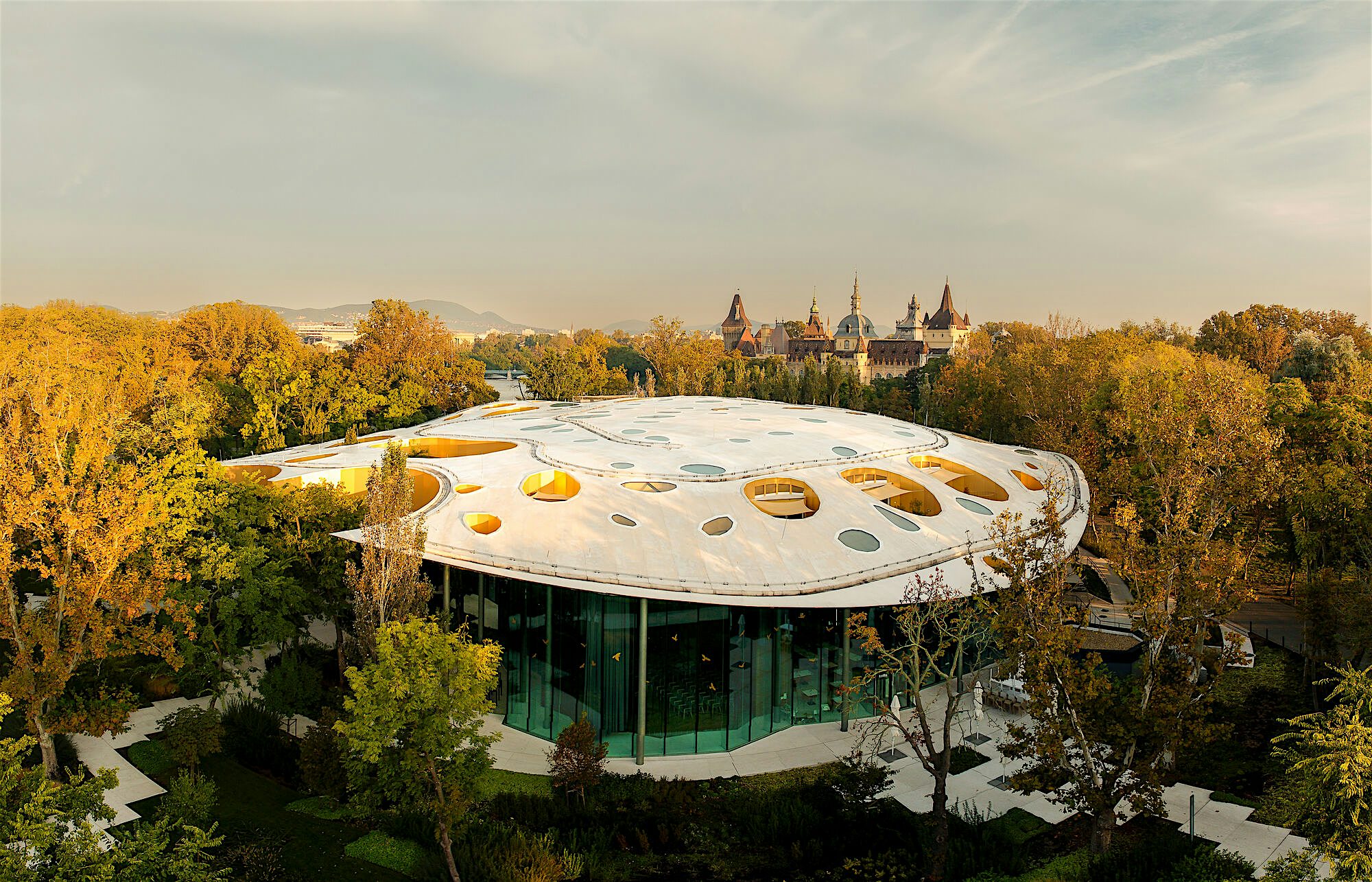 Modern circular pavilion with a white, oval roof punctuated by skylight openings, set among autumn‑coloured trees, with glass walls below and historic castle towers