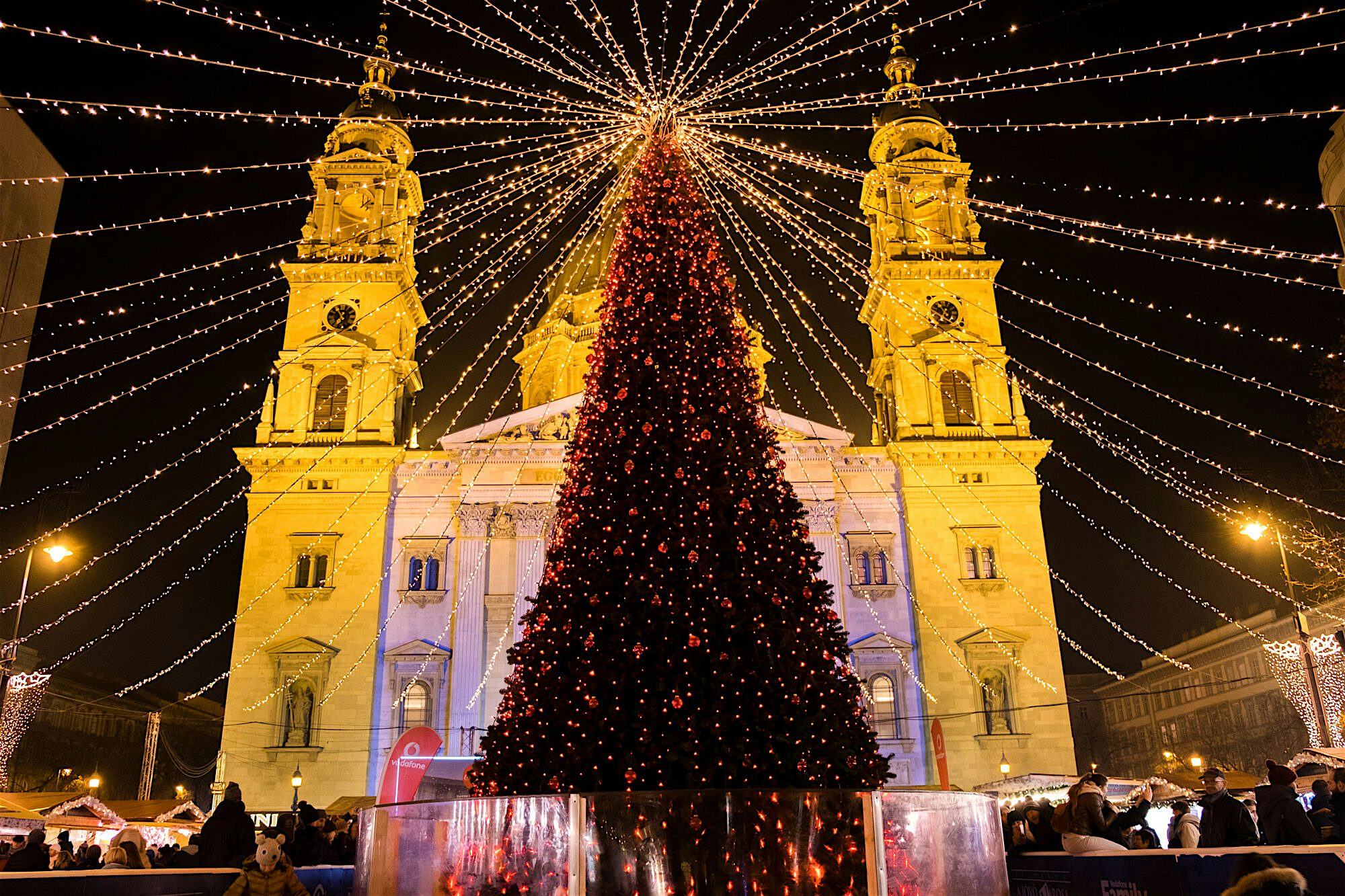 Large Christmas tree illuminated with festive lights in a city square, framed by strings of golden lights radiating overhead, with an ornate twin‑towered cathedral