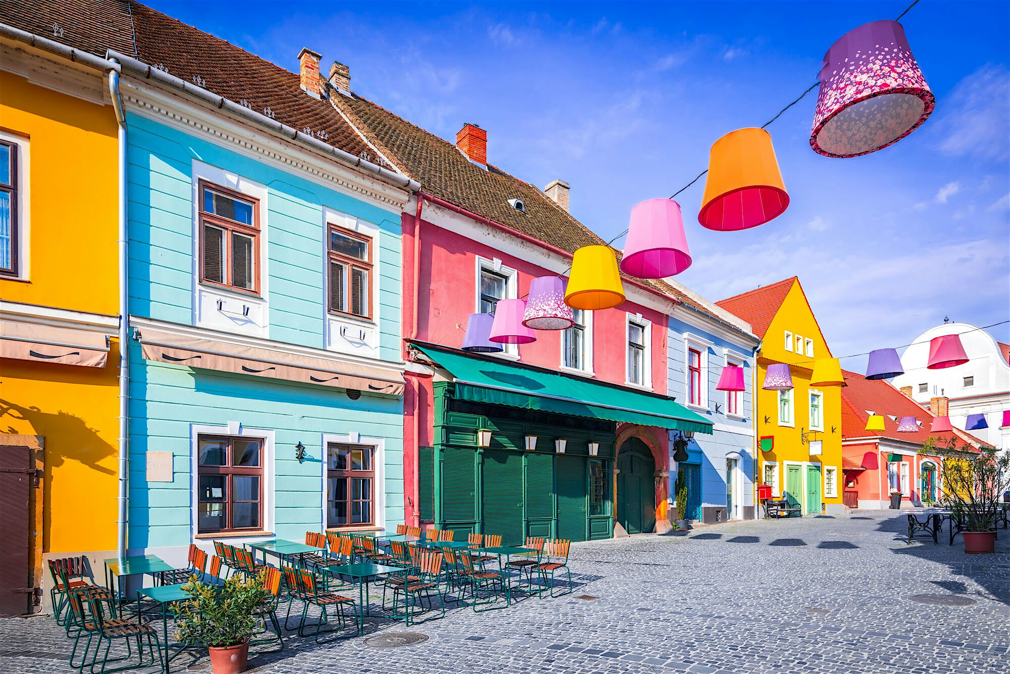 Colourful row of historic houses lining a cobbled square, painted in bright blues, yellows, pinks, and reds, with café tables outside and decorative lamps hanging overhead