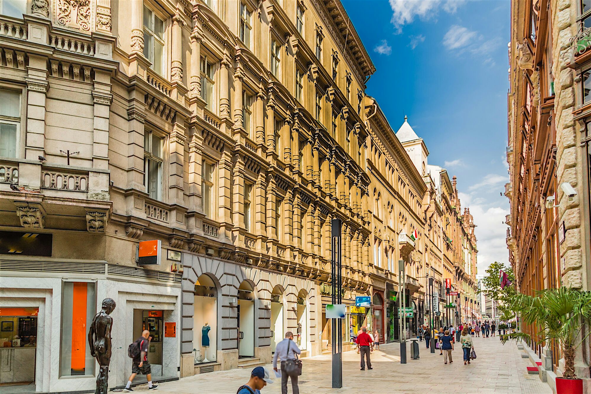 Street lined with ornate historic buildings featuring detailed stone facades, with pedestrians walking past shopfronts