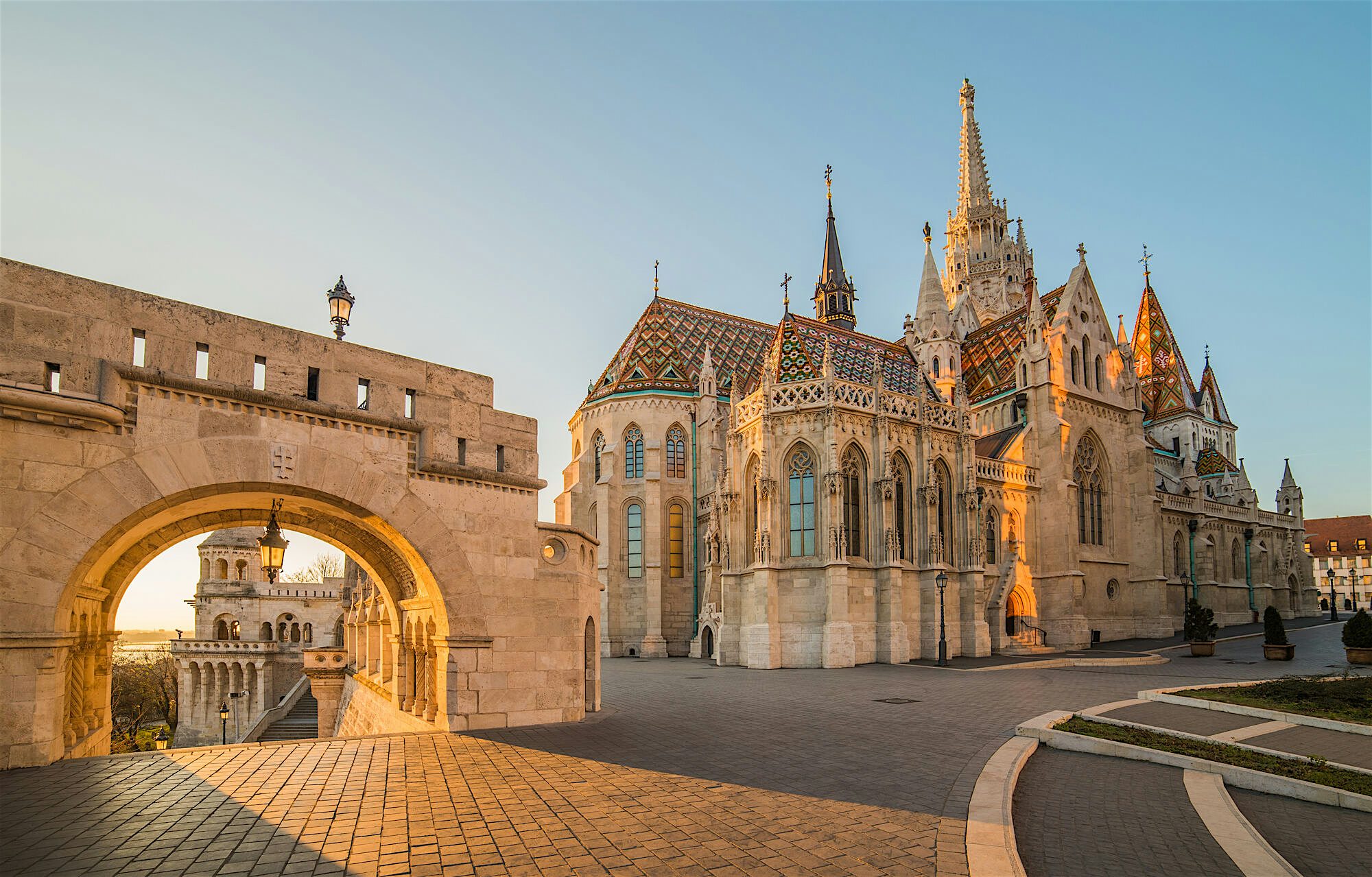 Sunlit historic church with patterned tiled roofs and tall spires, viewed from a stone terrace beside an arched gateway