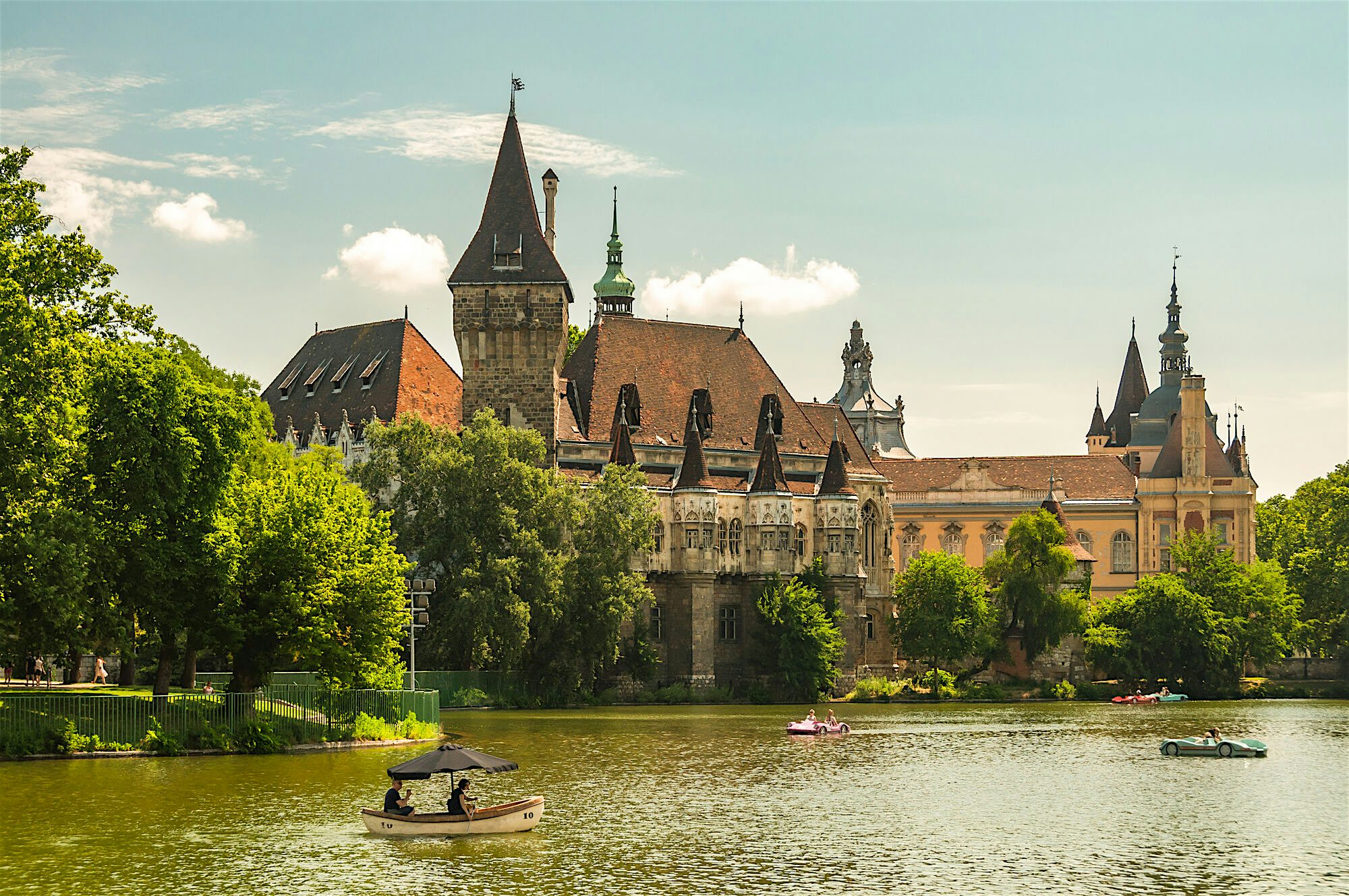 Historic castle‑style buildings with towers and ornate details rise behind a tree‑lined lake, with small boats
