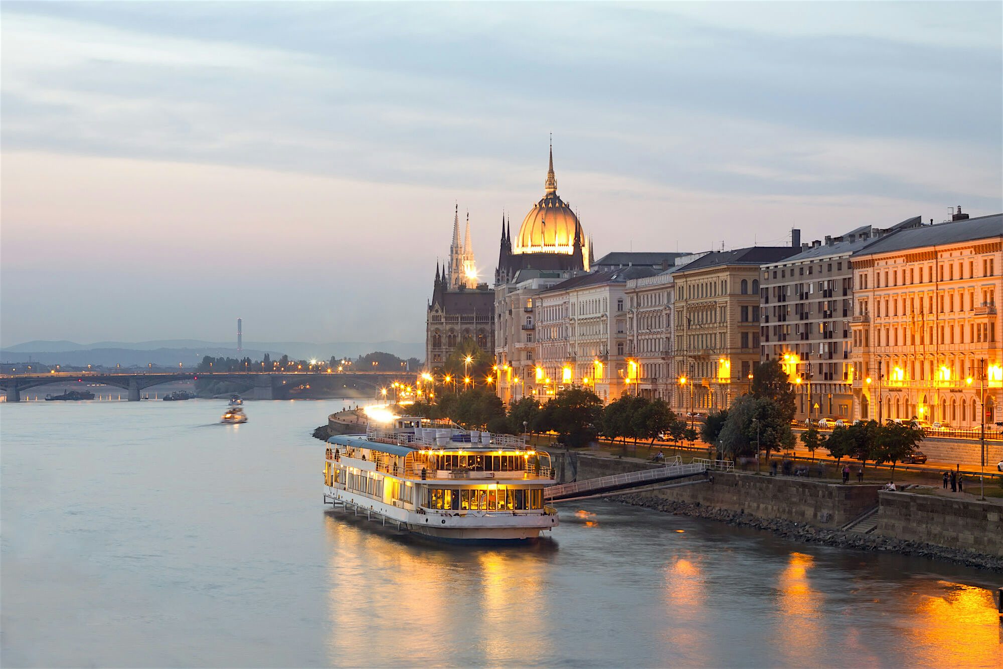 Riverside cityscape at dusk with illuminated historic buildings and a domed landmark along the waterfront, with a lit river cruise boat