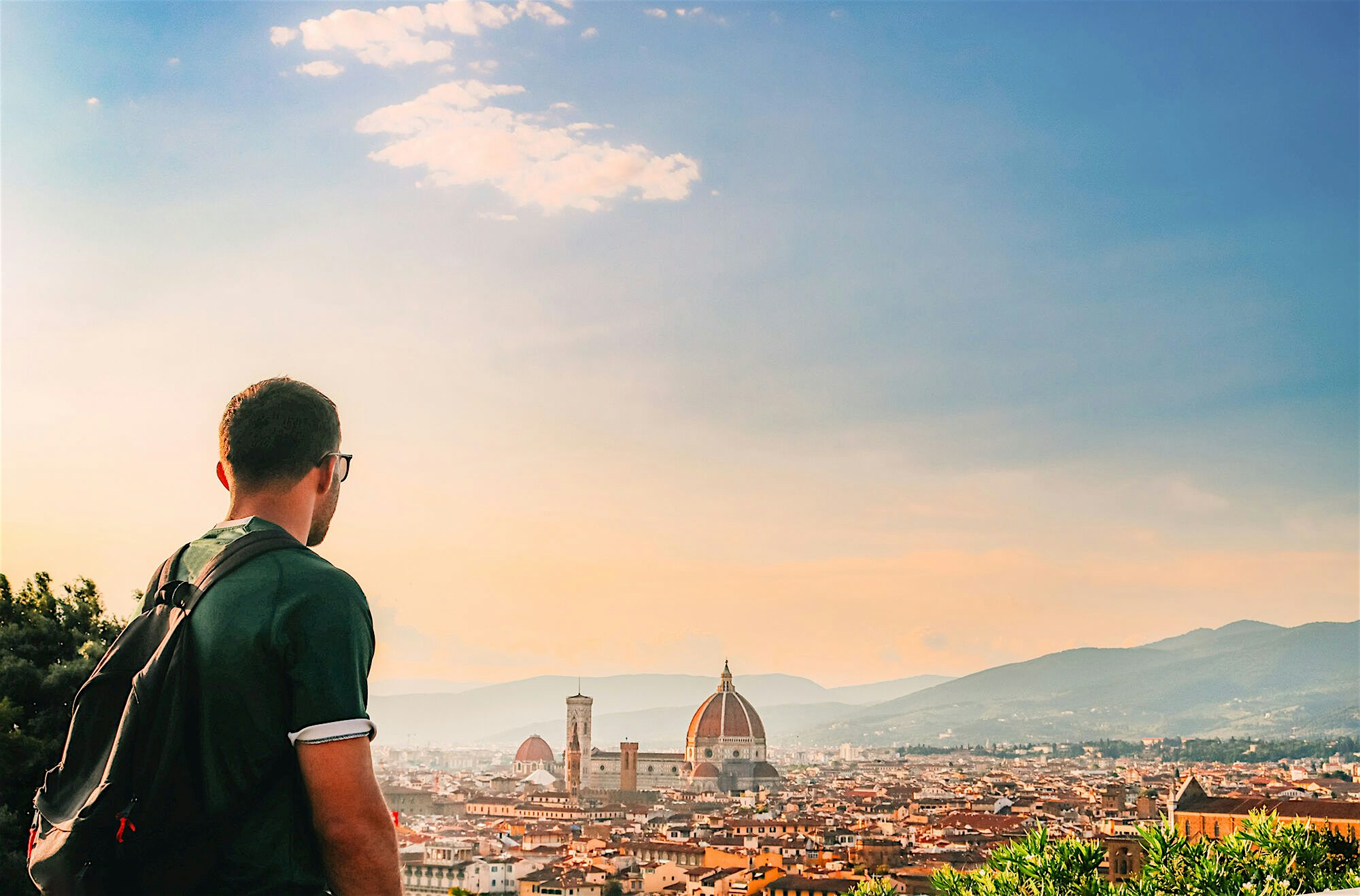 A person with a backpack overlooking a historic city skyline, with a large domed cathedral rising above the rooftops in the distance.