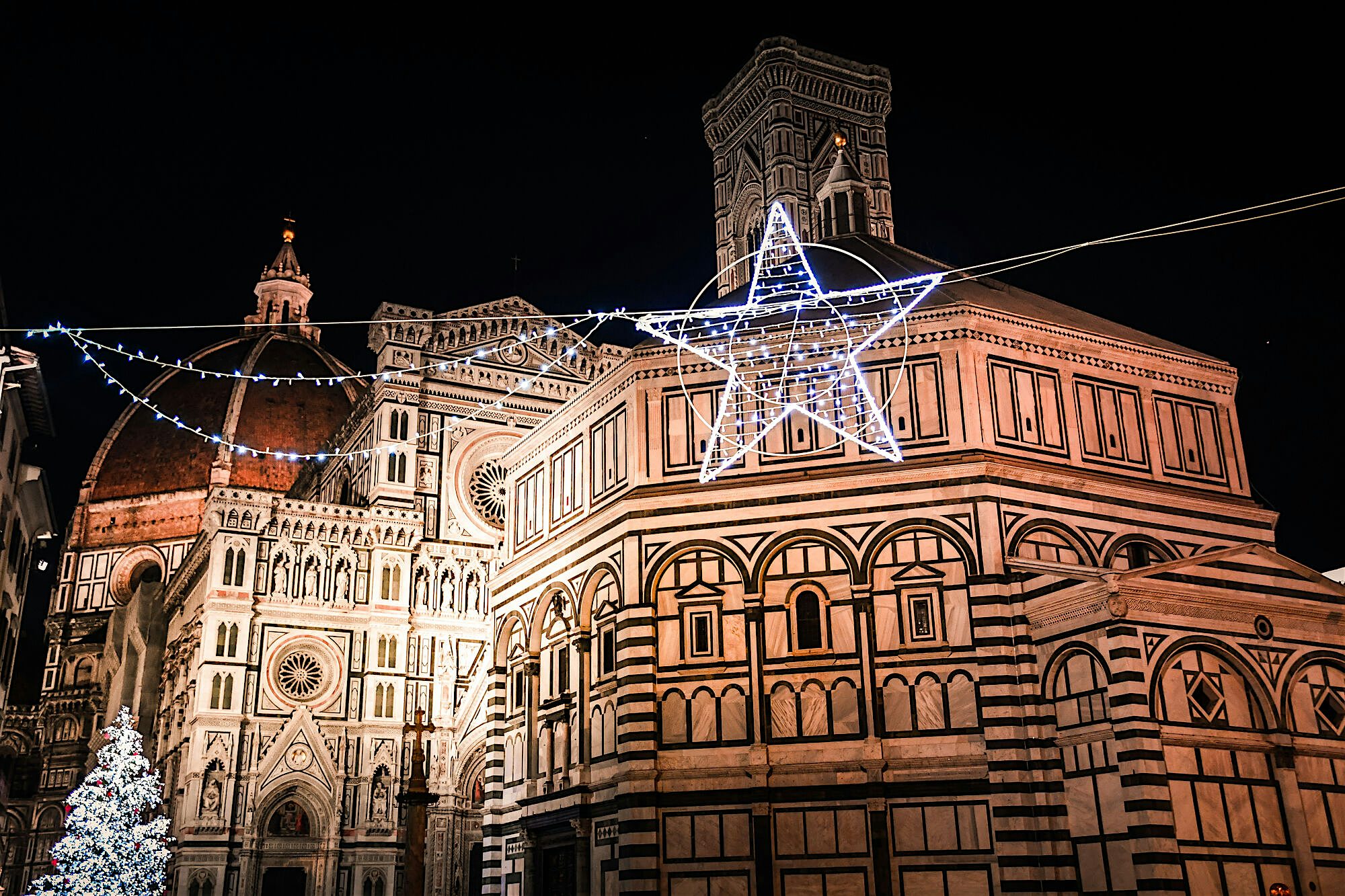 "An illuminated historic cathedral and domed buildings at night, decorated with festive lights and a large star suspended above the square.  "
