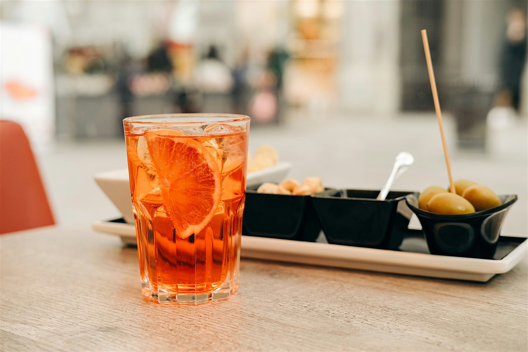 An orange drink in a glass with ice and a fresh slice of orange, with olives and other sides in a dish behind