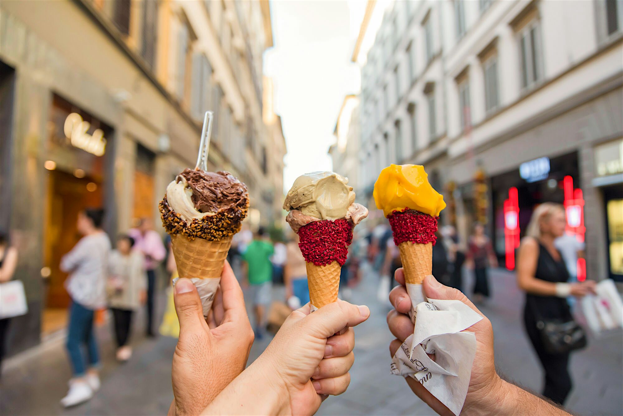Three icecreams being held infront of the camera, assorted flavours and sprinkles around the waffle cones. An old shopping district in the background