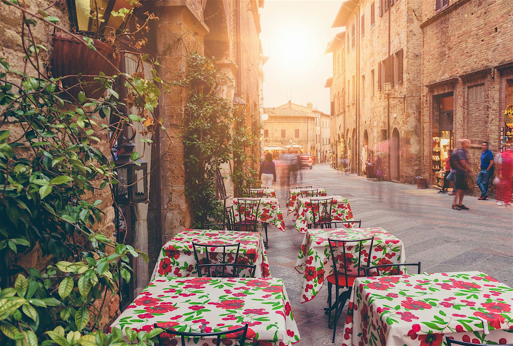 "Outdoor café tables with patterned tablecloths lining a narrow cobbled street, with historic stone buildings and warm evening light.  "