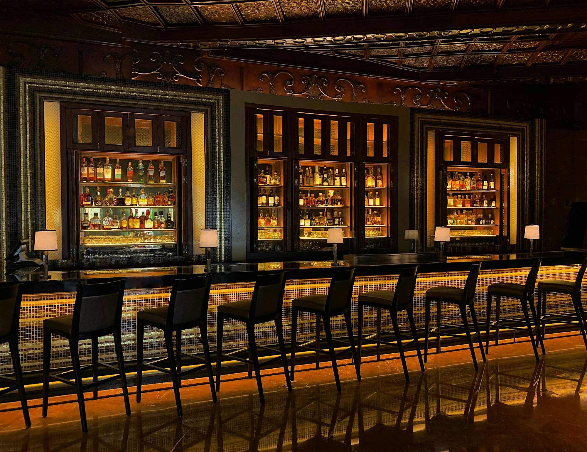 A dark bar with a line of bar stools lining it, ornately decorated and back lighting of the liquor cabinets and coming up beneath the bar