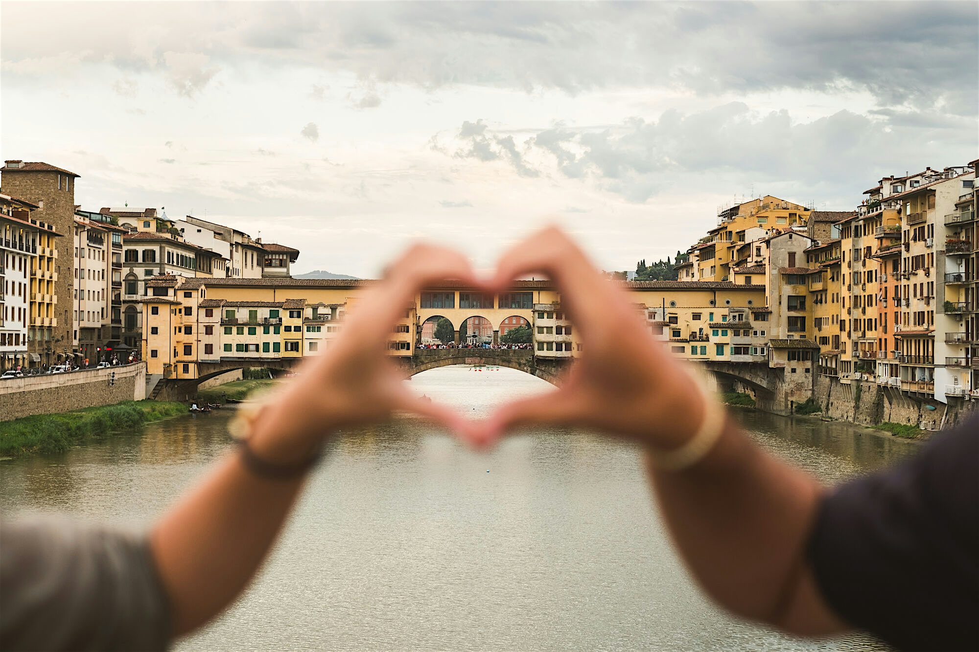 Two hands forming a heart in front of the camera, behind you can see the river and Ponte Vecchio, Florence's oldest medieval stone bridge