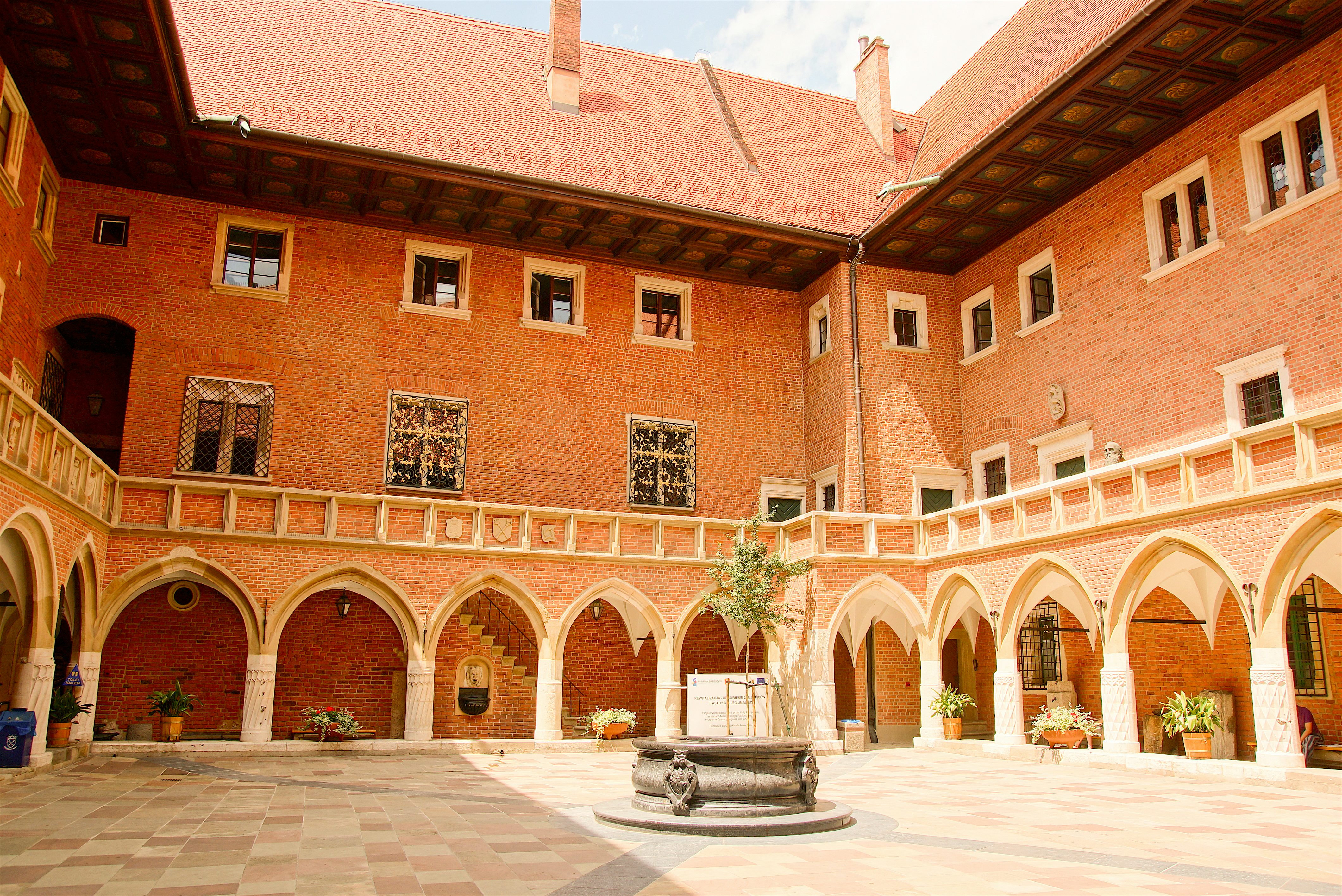 Enclosed courtyard with red brick buildings and arched colonnades on all sides, a central stone well, patterned paving, and small windows and decorative details