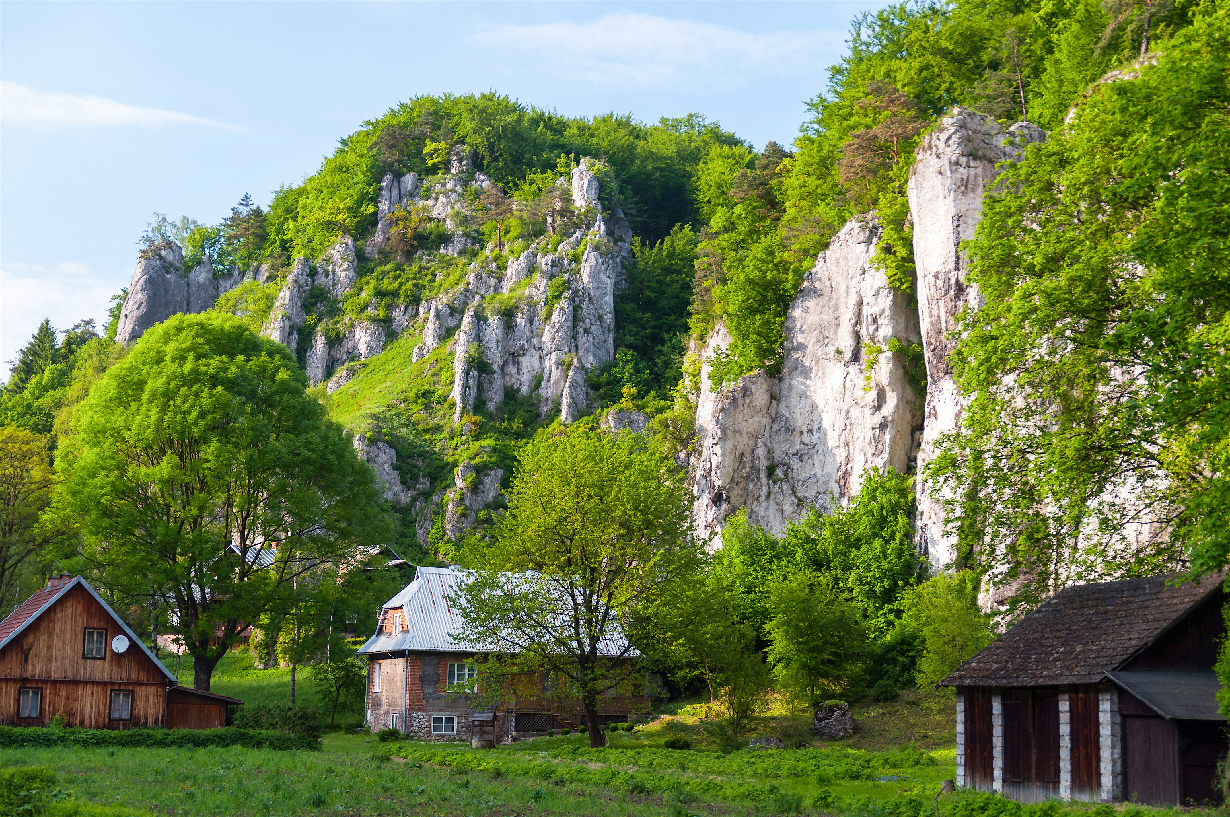 Small rural houses set in a green valley beneath steep limestone cliffs, with trees and grassy meadows in the foreground and rocky outcrops