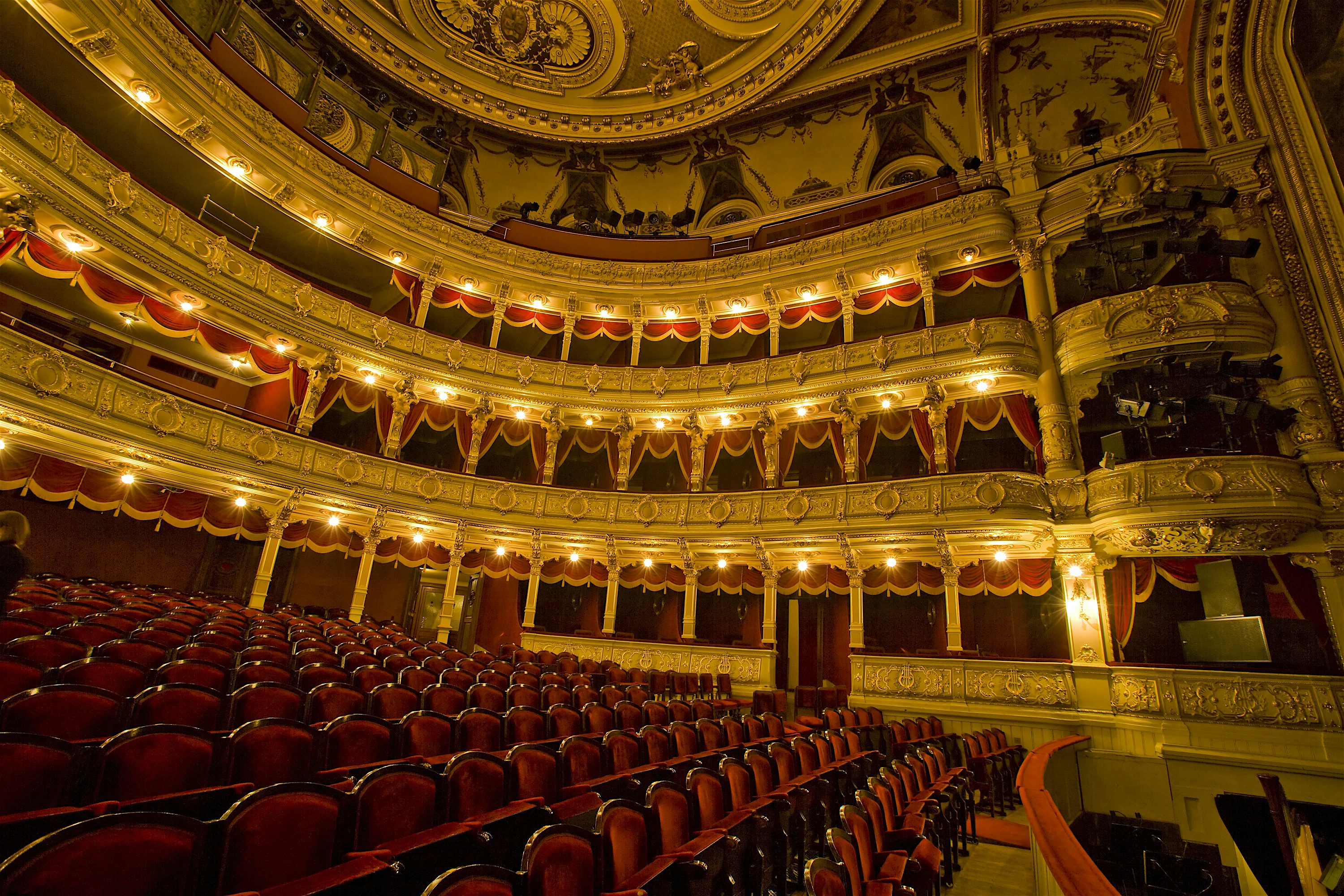 Ornate theatre interior with rows of red velvet seats facing an empty stage, surrounded by multiple tiers of gilded balconies, decorative arches