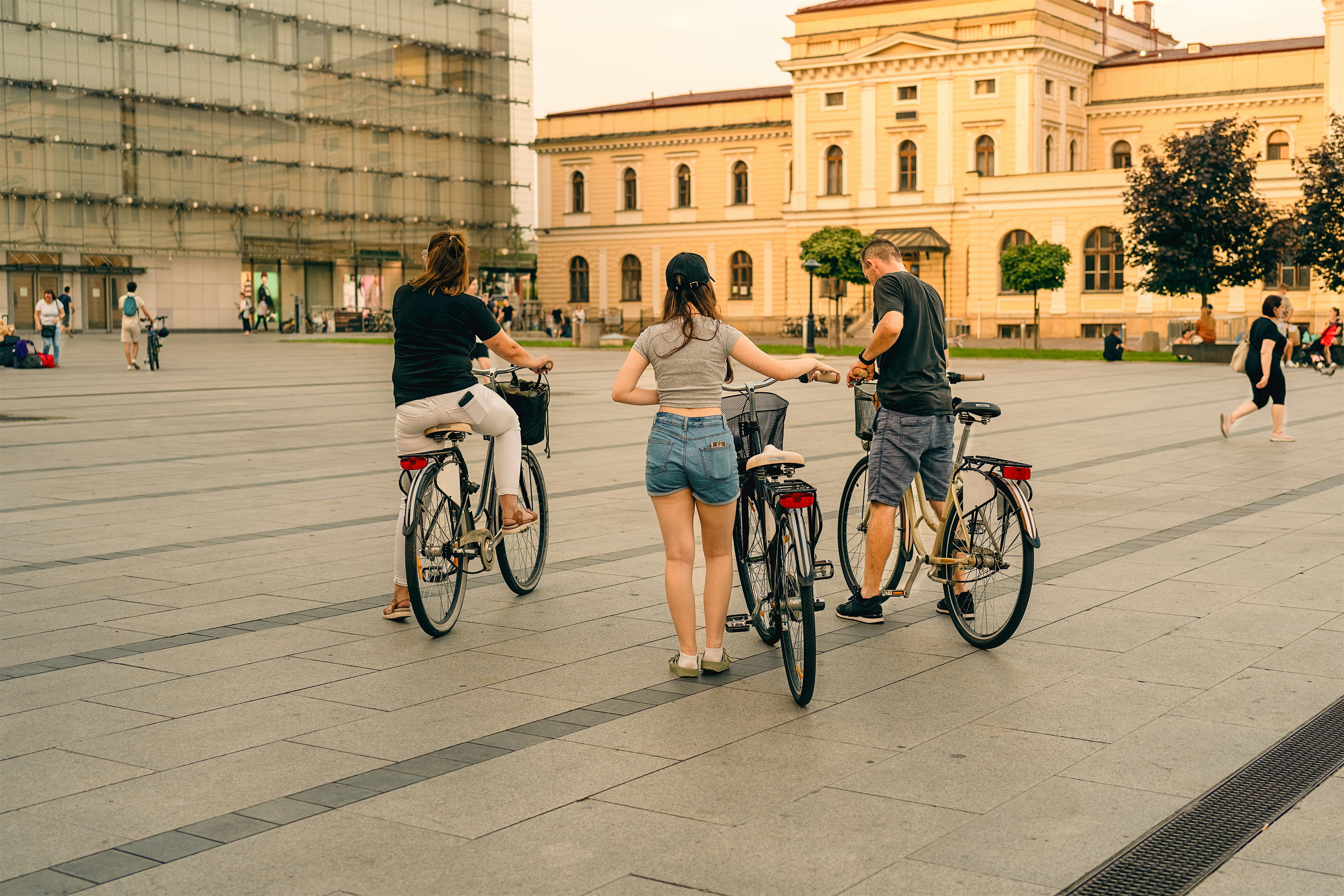 Three people standing with bicycles in a wide urban square, with historic buildings and trees