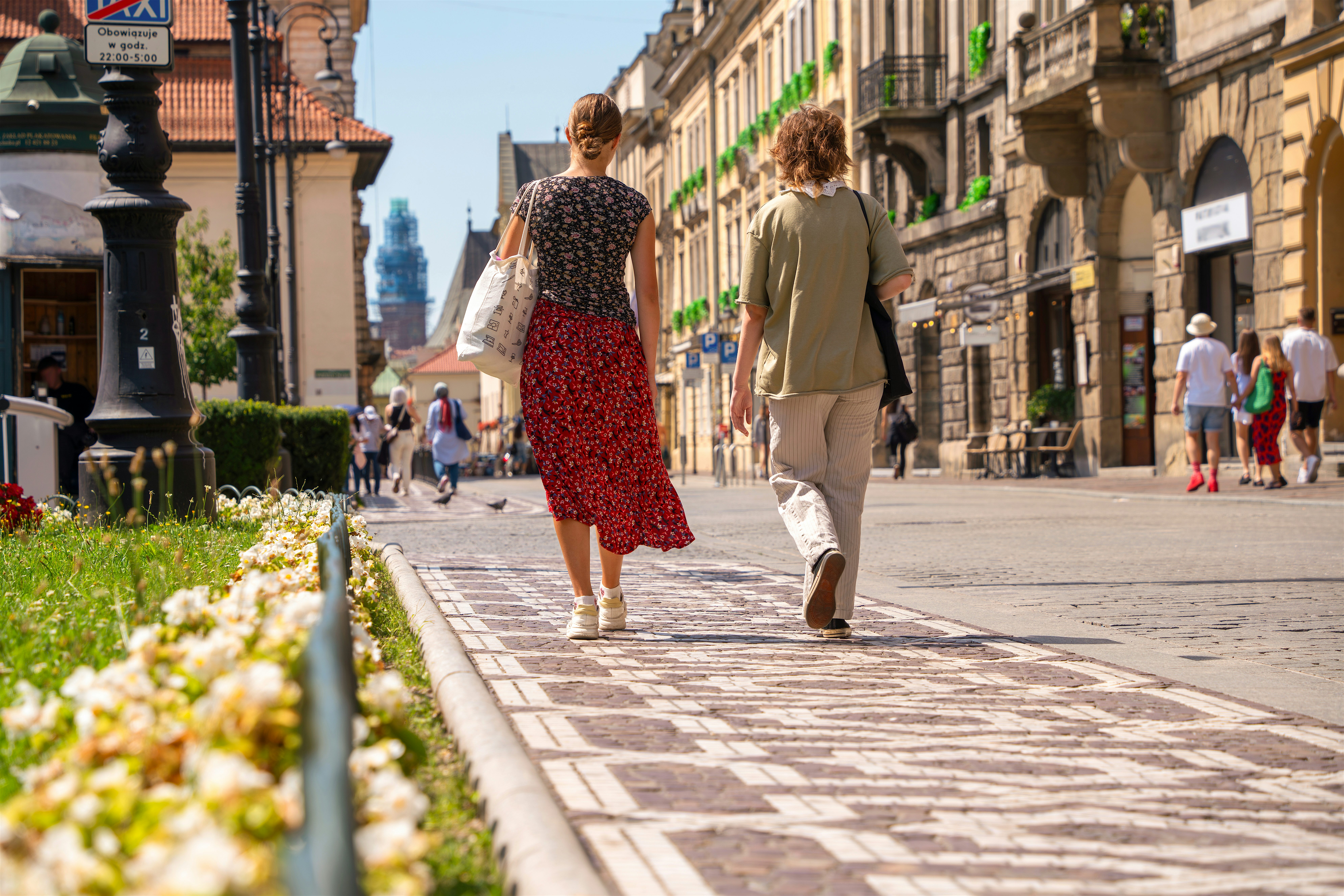 Two people walking along a sunlit city street lined with historic buildings and shopfronts, with flowers