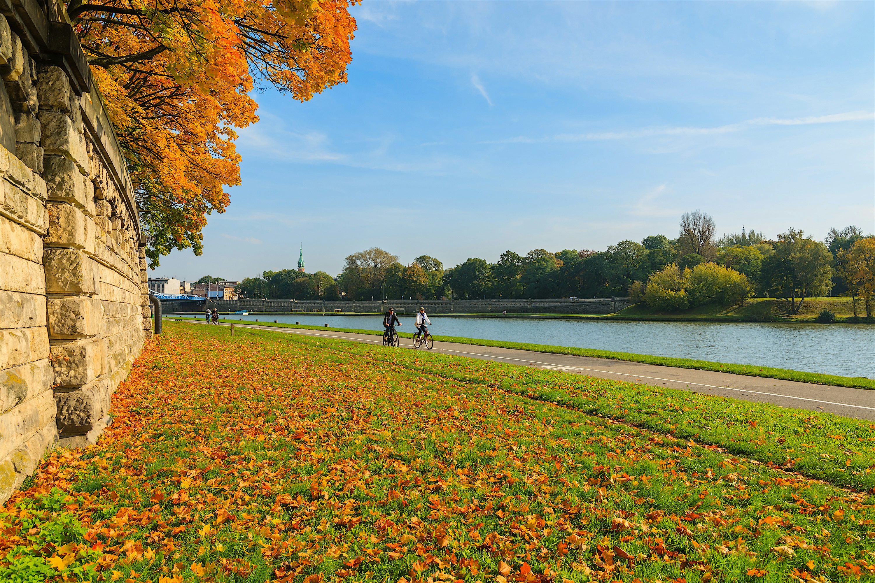 Leaf‑covered riverside path in autumn, with golden trees along a stone wall, people cycling beside calm water