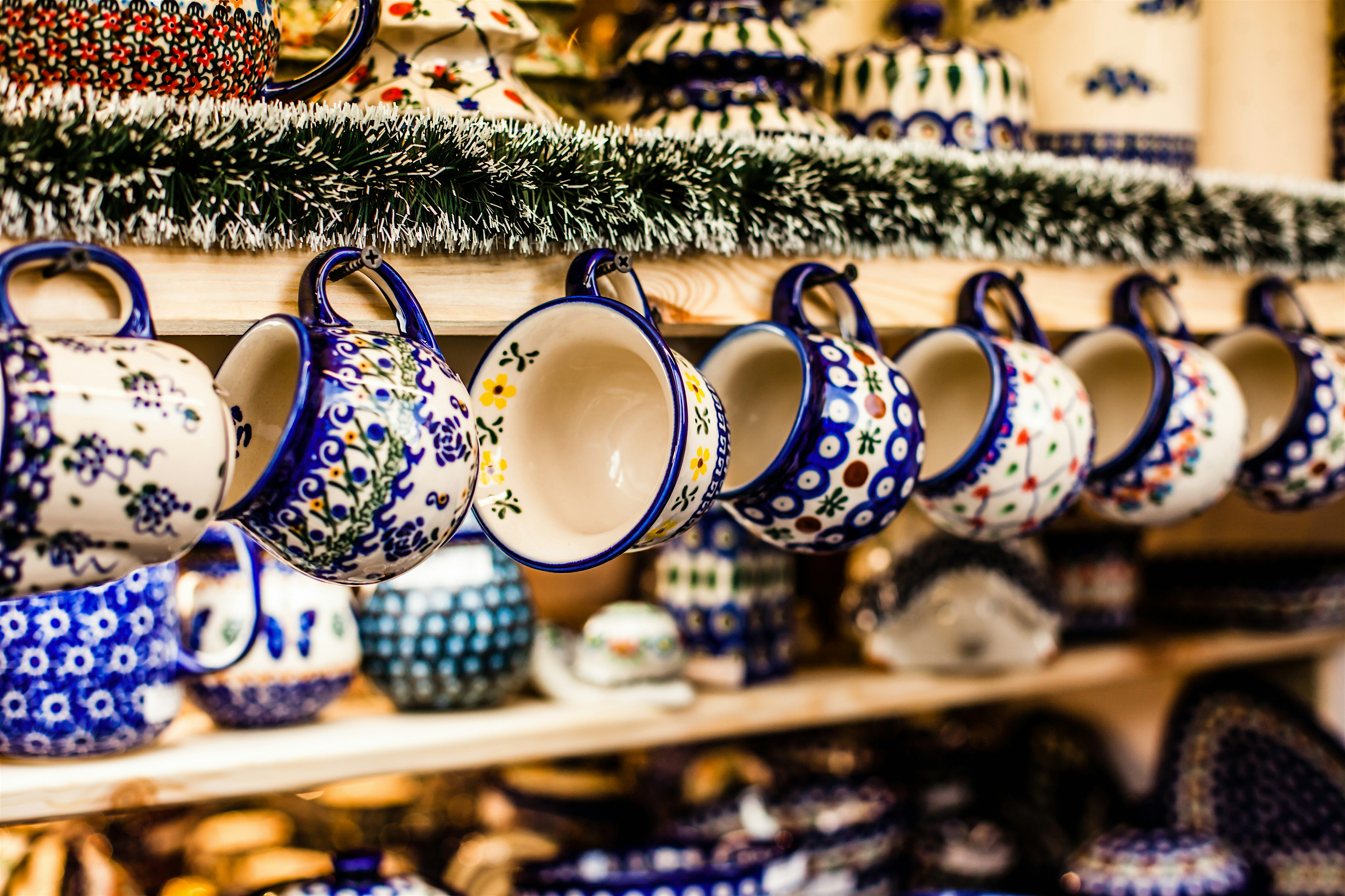 Row of patterned ceramic mugs hanging from a wooden shelf, decorated with intricate blue, green, and red designs, with more matching pottery arranged below.