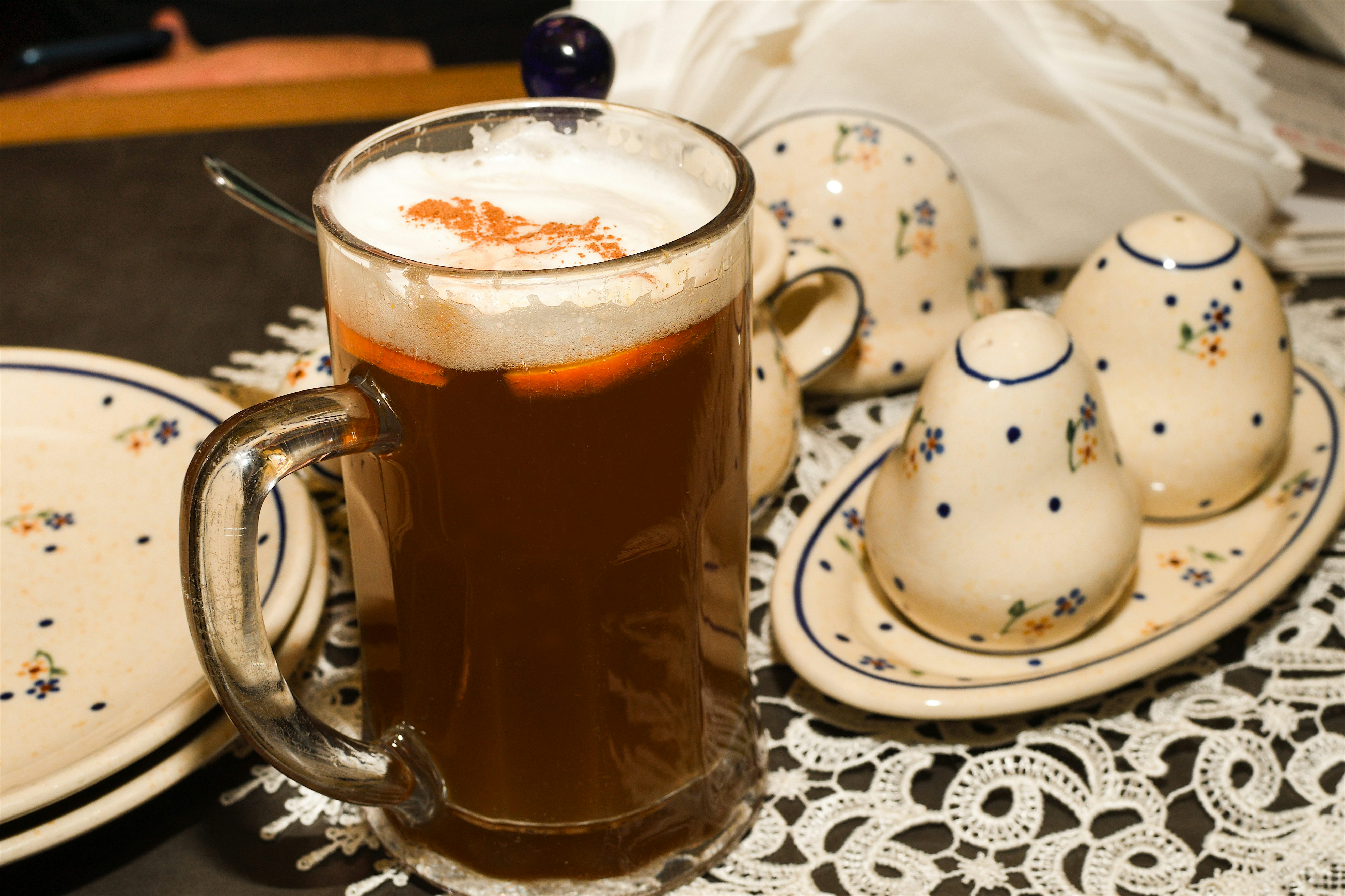 Glass mug of dark beer topped with a foamy head, served on a lace tablecloth with small ceramic cups