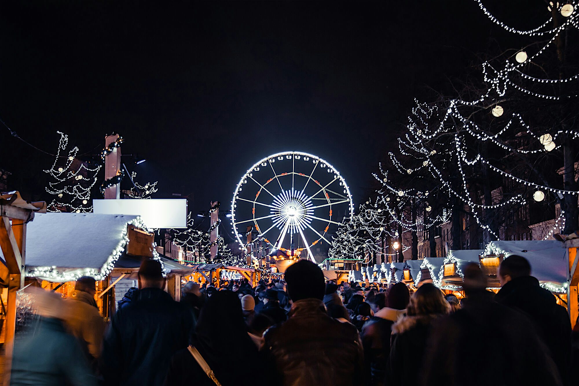 Crowded night market filled with festive lights and wooden stalls, with a large illuminated ferris wheel rising above the crowd.