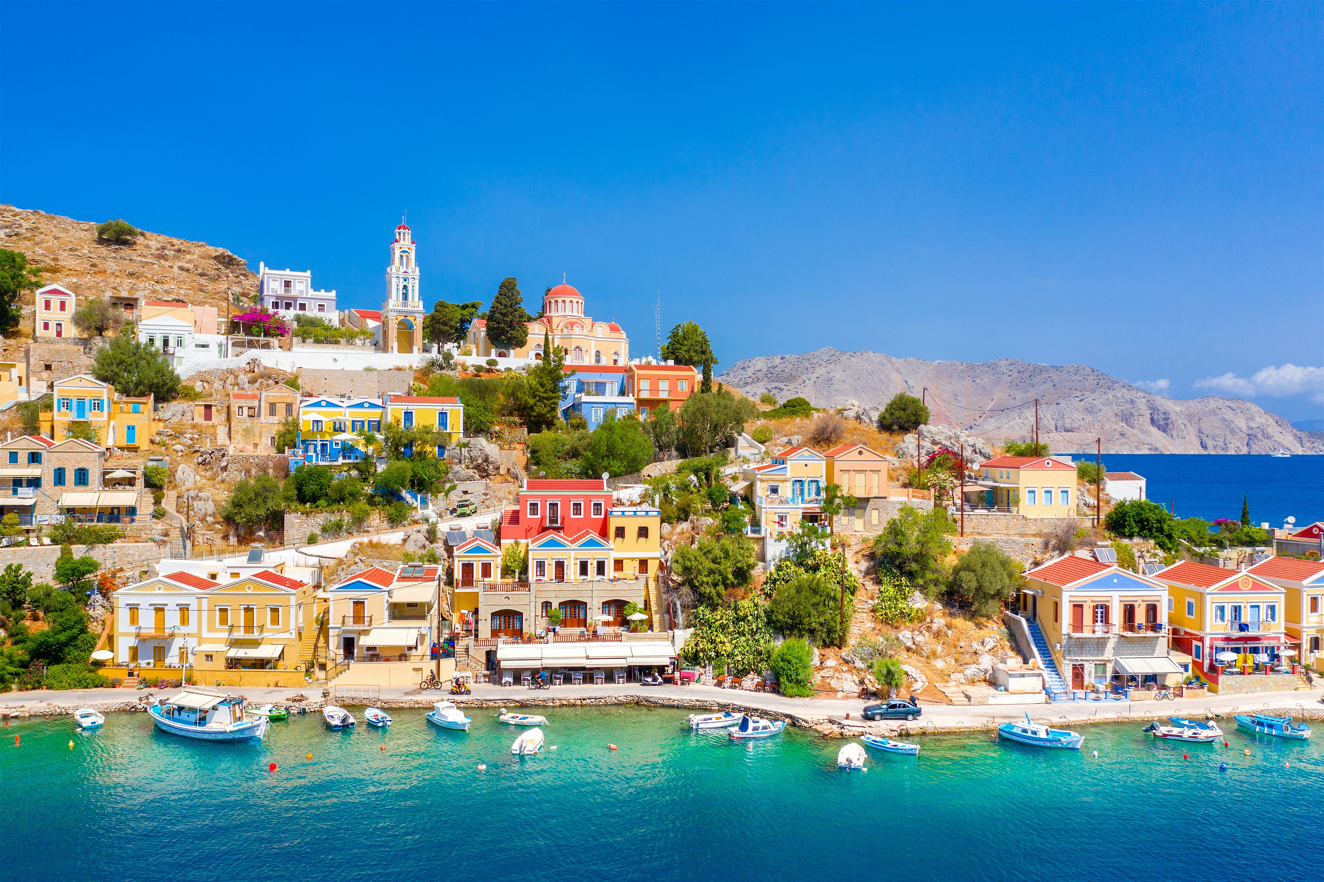 A colourful hillside town overlooks clear blue water, with small boats bobbing in the harbour below rows of vibrant houses and a prominent church rising above the skyline.