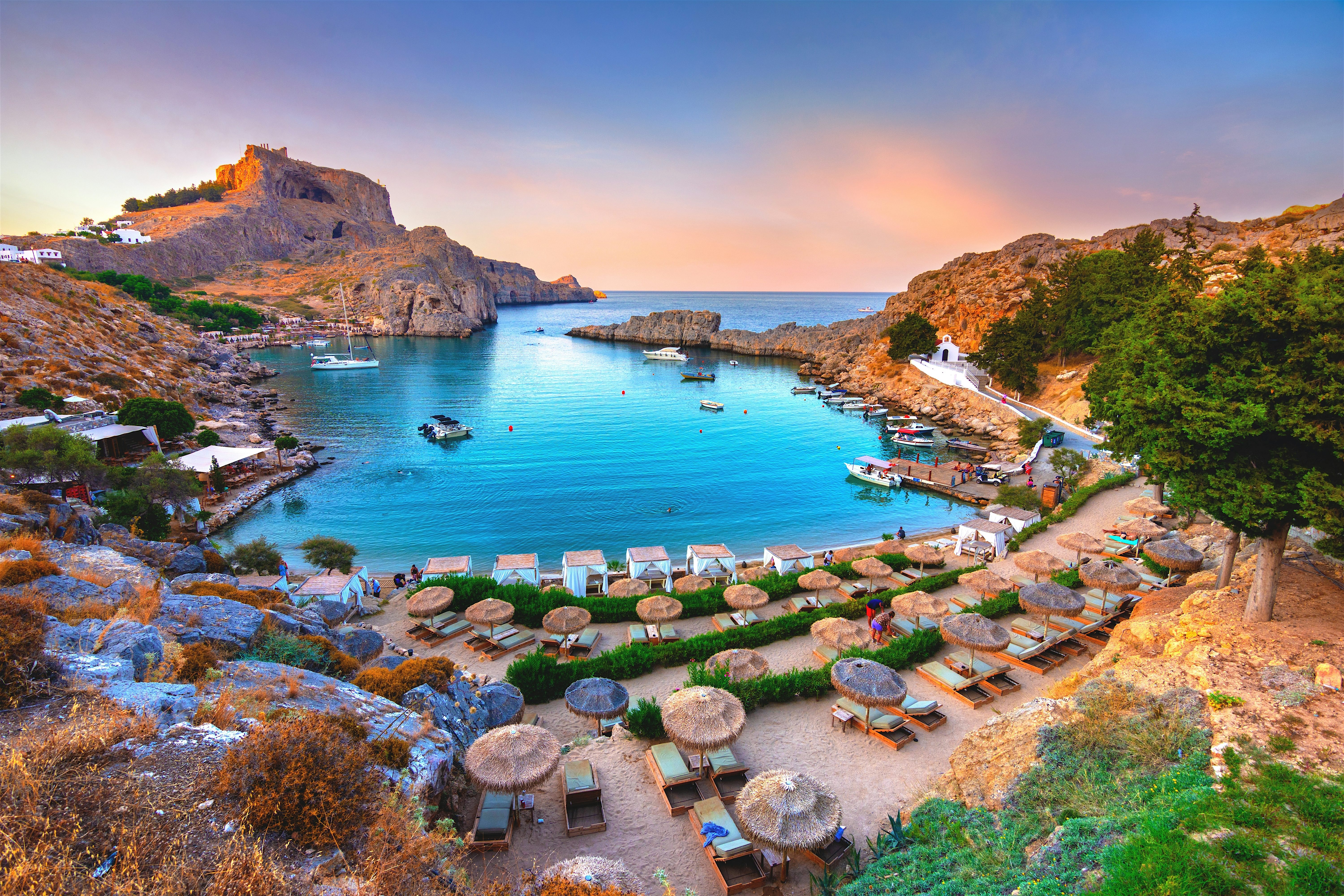 A sheltered turquoise bay curves between rocky cliffs, with boats on the water and sun loungers lined along the shore below.