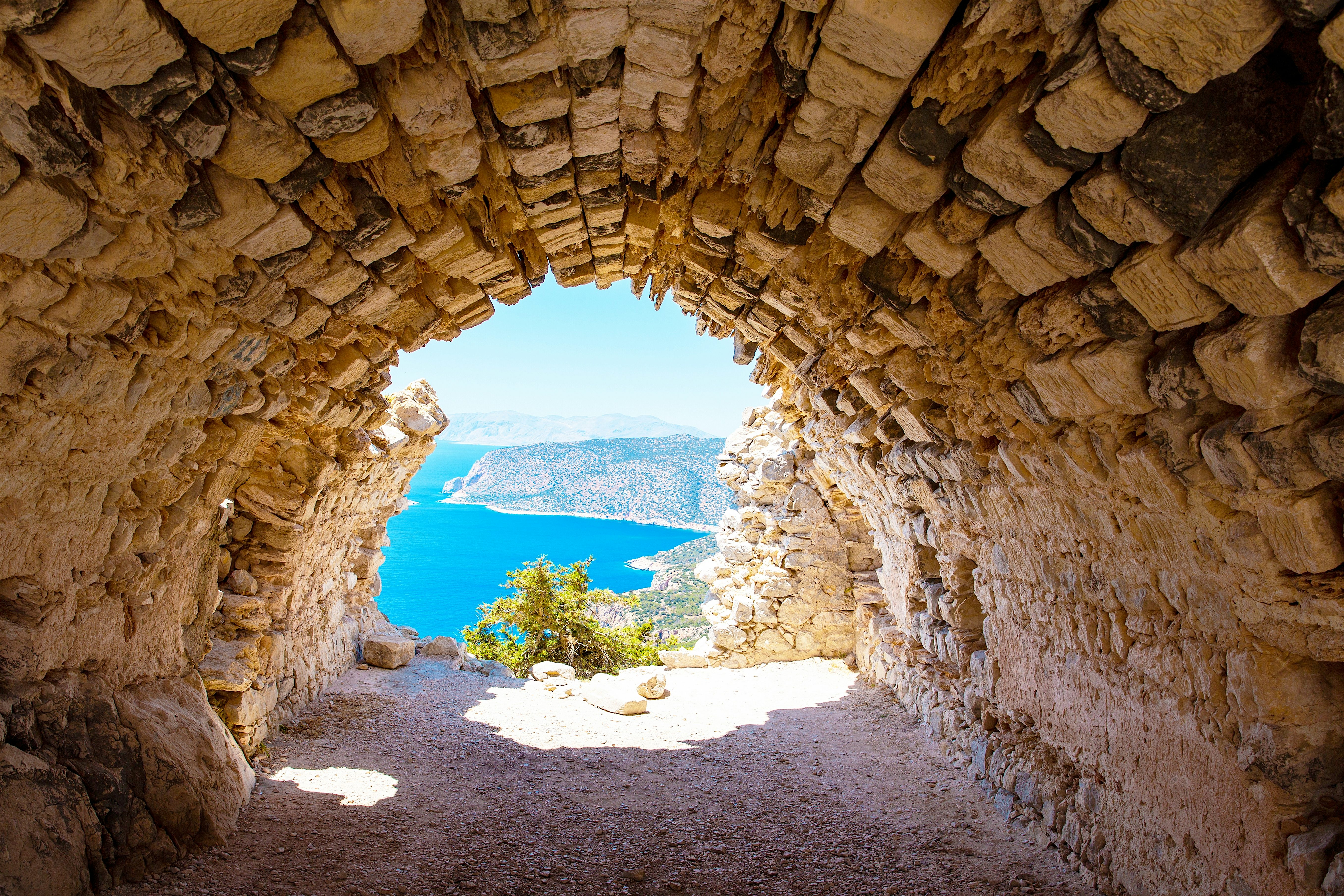 A rugged stone arch frames a bright coastal view, opening out onto vivid blue sea and distant hills beyond a sunlit clearing.