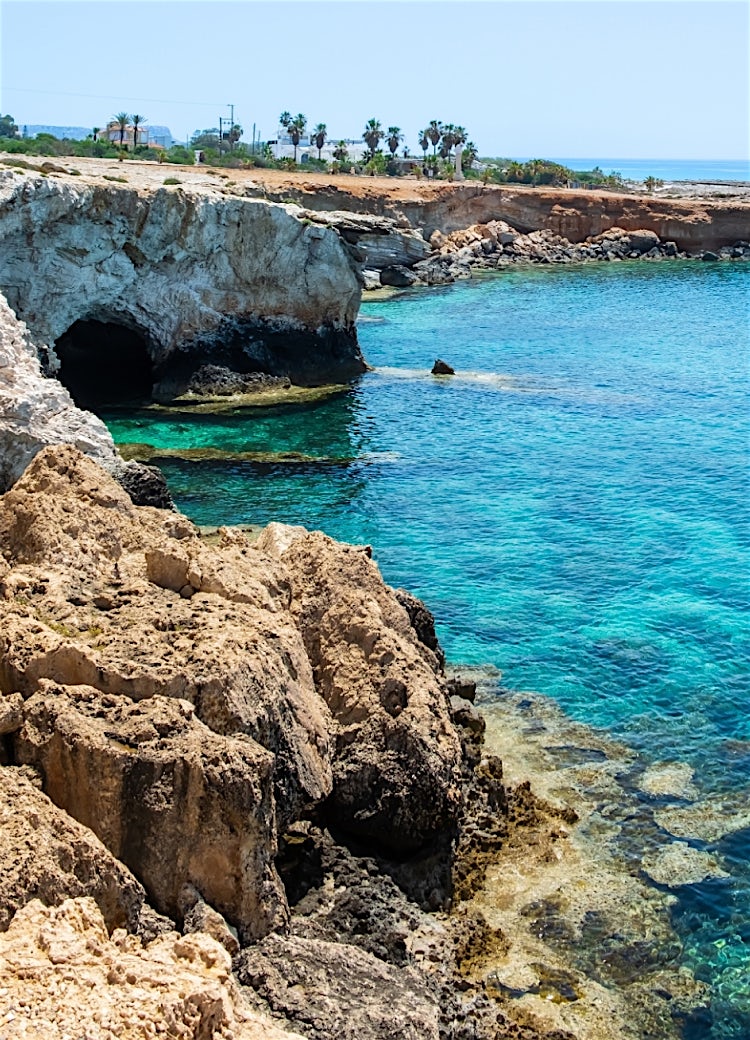 Crystal-clear turquoise waters surrounding coastal rock formations and a natural sea arch along a Mediterranean shoreline in Cyprus.