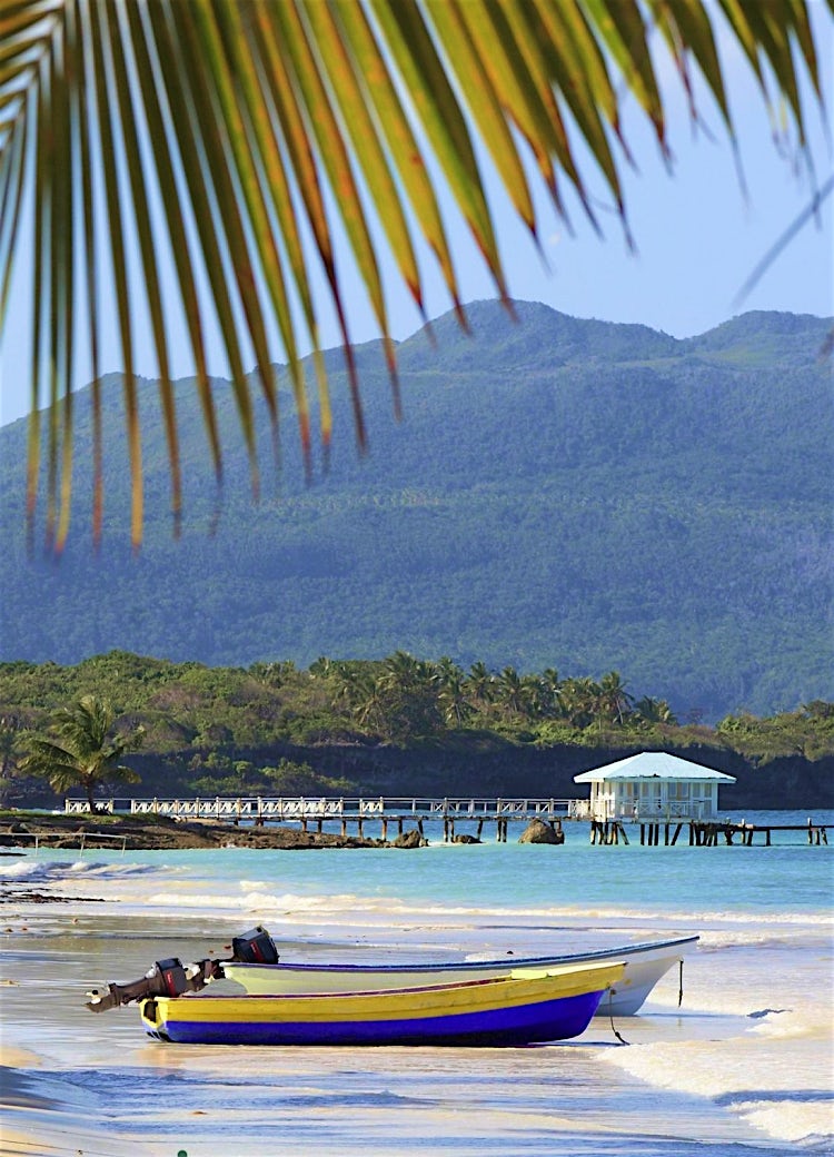 Dominican Republic tropical beach with fishing boats, palm-fringed shoreline, turquoise sea and lush coastal mountains.