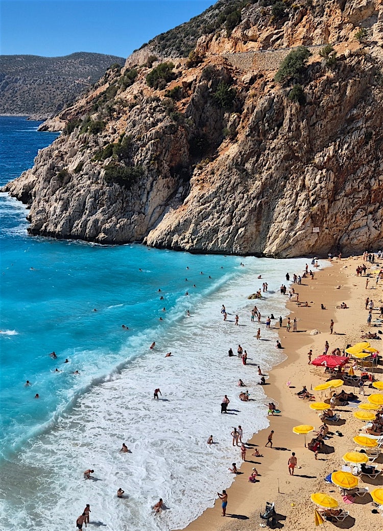 Turkish beach cove with turquoise Mediterranean waters, golden sand, dramatic cliffs and sun umbrellas along the coastline.