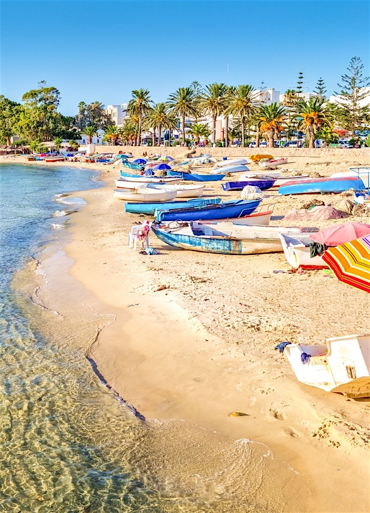 Tunisia beach with traditional fishing boats, palm-lined shoreline, clear Mediterranean waters and scenic coastal resort town.
