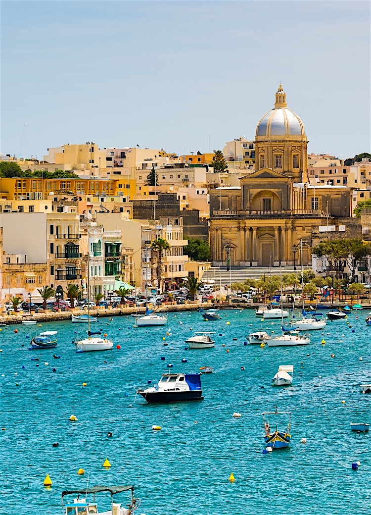 Malta waterfront harbour with historic church, moored boats, turquoise waters and traditional Mediterranean architecture.