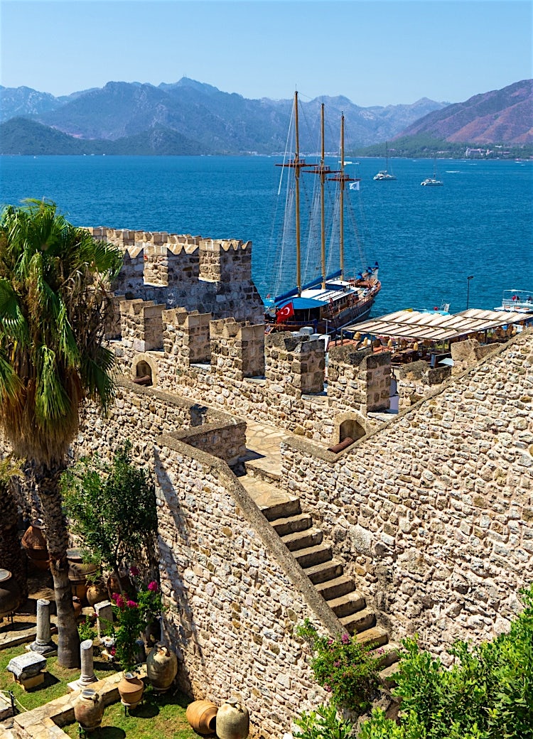 Historic Marmaris Castle overlooking the turquoise Aegean coast, with traditional sailing boat, marina and mountain backdrop, Turkey.