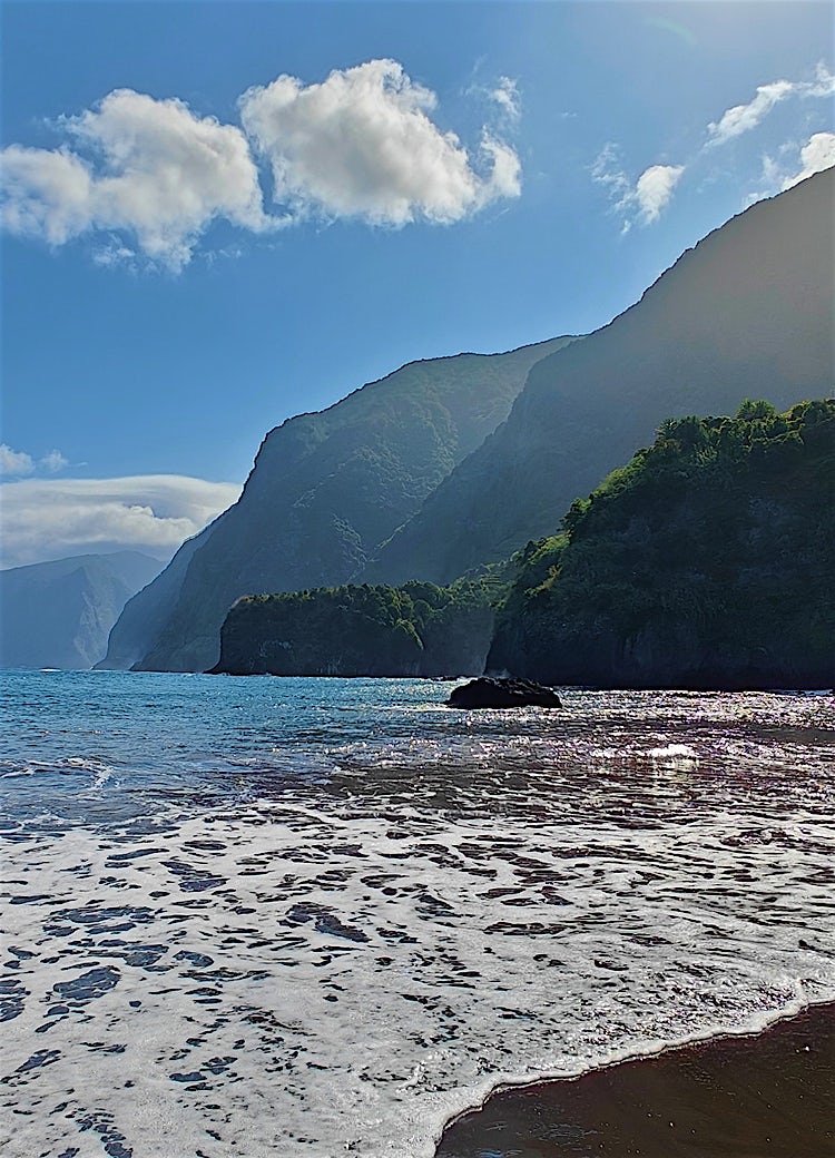 Madeira coastline with dramatic volcanic cliffs, black sand beach, Atlantic Ocean waves, and lush green mountain scenery.