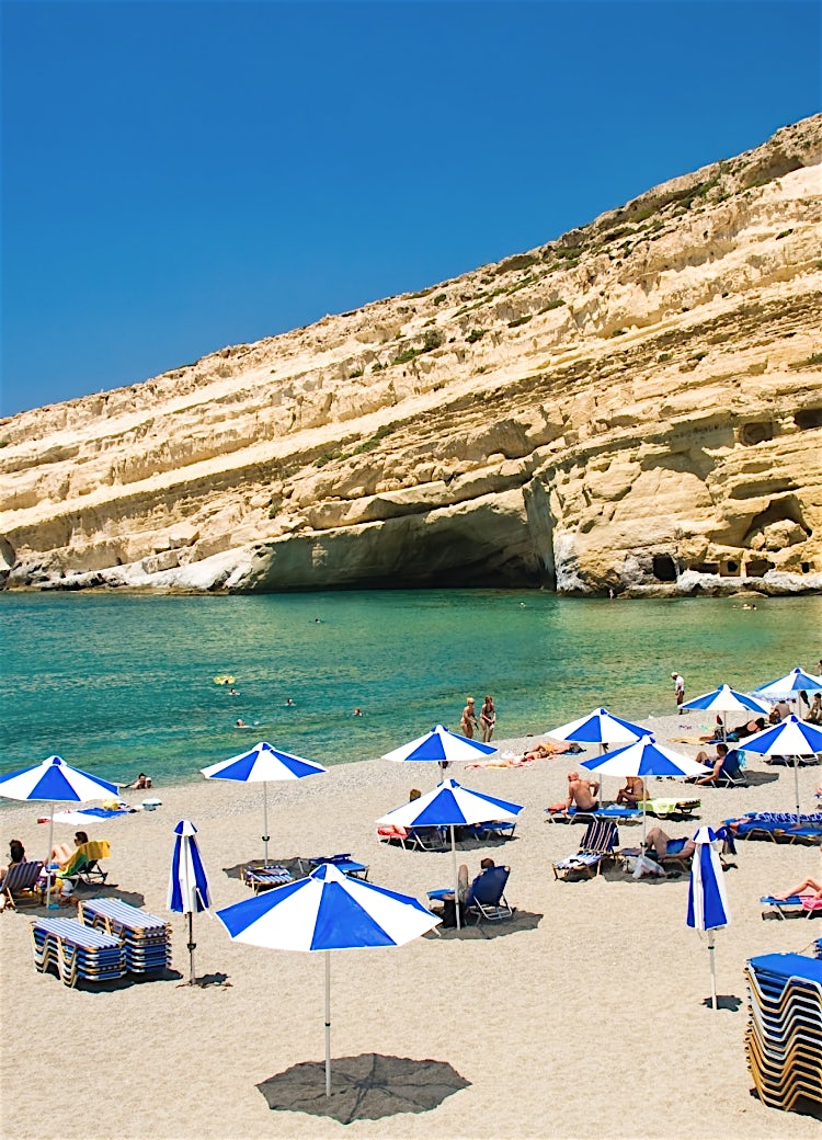 Matala Beach, Crete, Greece, with turquoise waters, seaside caves, sandy shoreline and sun umbrellas beneath dramatic coastal cliffs.