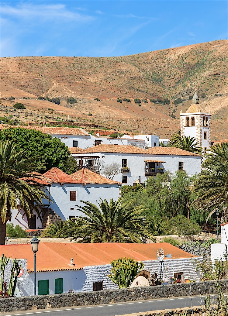 Fuerteventura village with whitewashed buildings, palm trees, historic church tower and rugged volcanic mountain scenery.