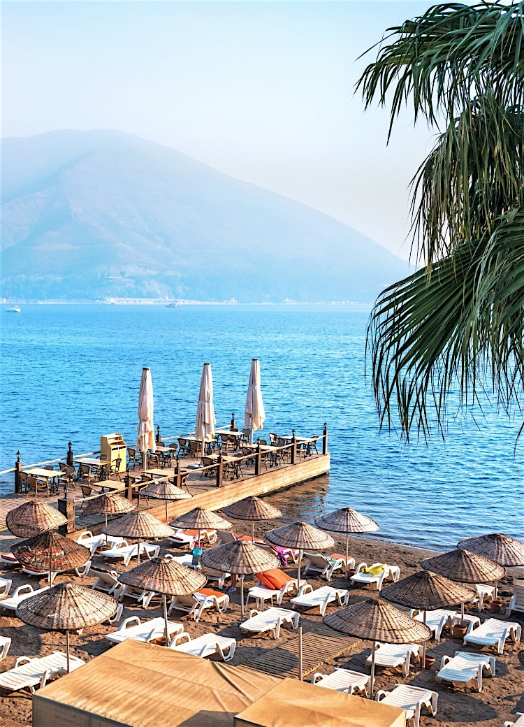 Tropical beachfront resort in Marmaris, Turkey, with sun loungers, parasols, wooden pier restaurant, calm blue sea and mountain backdrop.