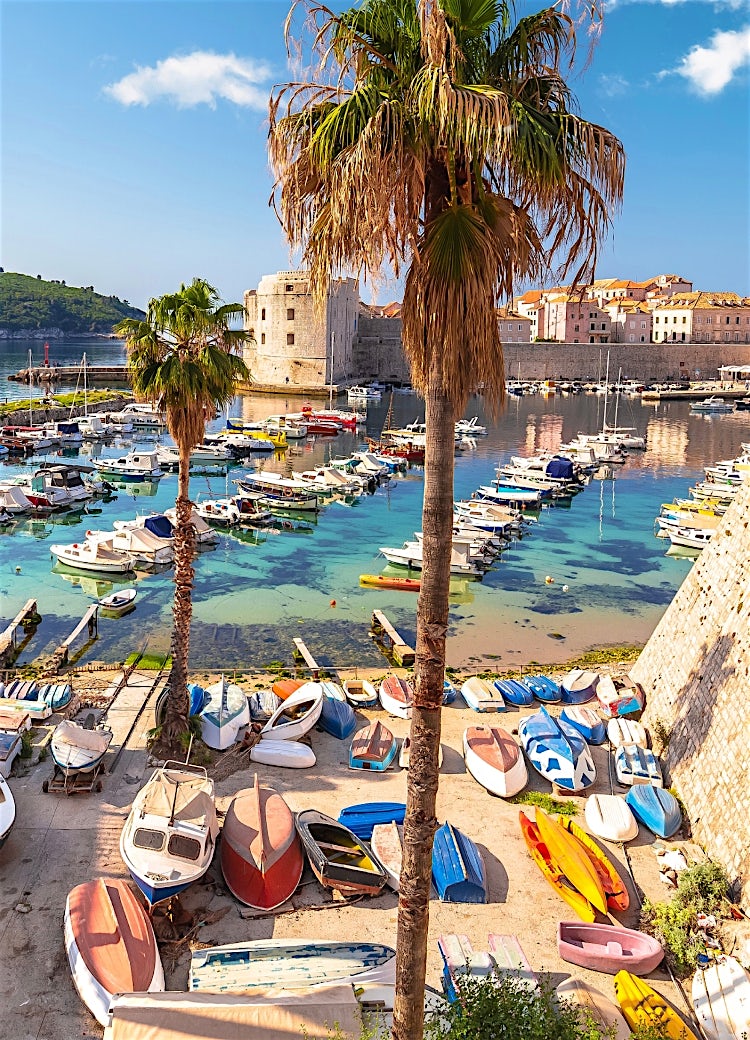 Dubrovnik Old Harbour, Croatia, with boats in crystal-clear Adriatic water, historic stone walls, palm trees, and a scenic coastal cityscape.