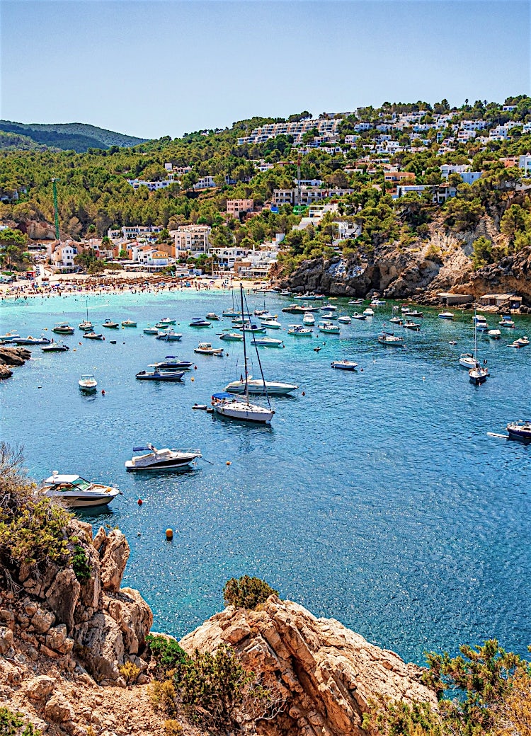 Ibiza beach cove with turquoise Mediterranean waters, anchored boats, rocky coastline, and hillside resorts overlooking a scenic bay.