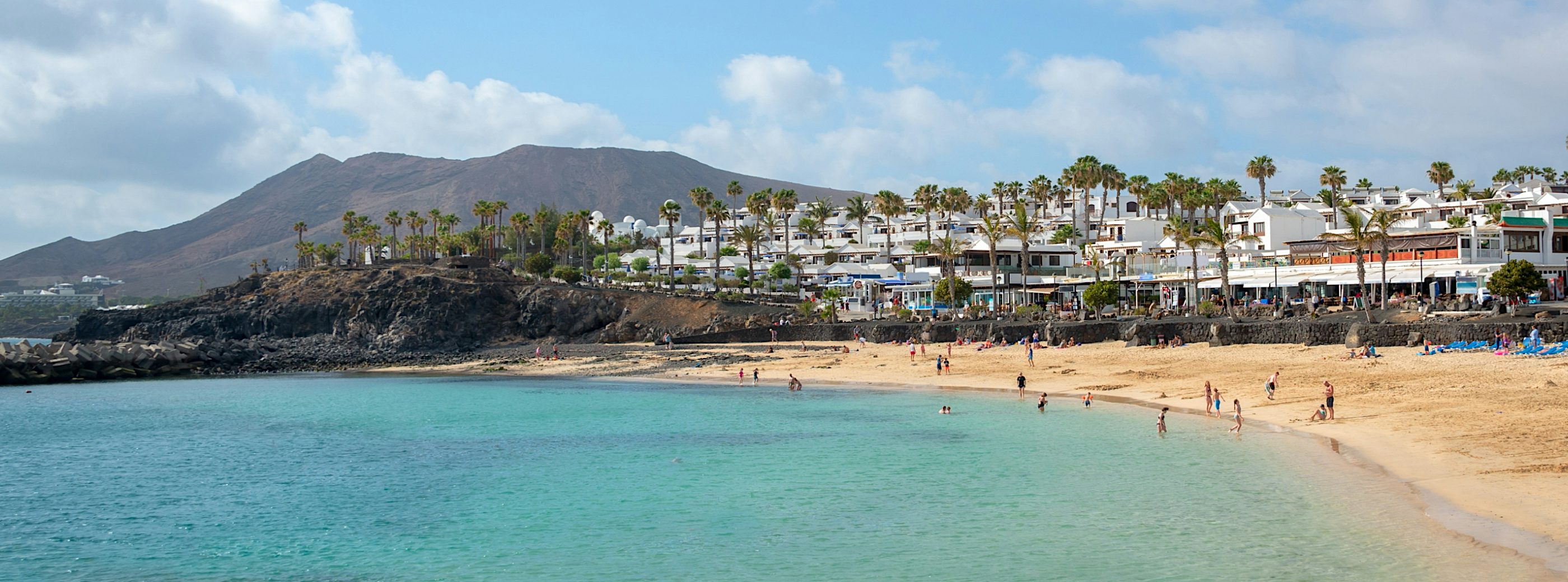 Turquoise-water beach in Lanzarote with palm-lined waterfront, whitewashed buildings and volcanic mountains in the background.