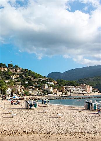 Palm-lined beach in Majorca with sandy shoreline, coastal promenade, hillside town and mountain backdrop.
