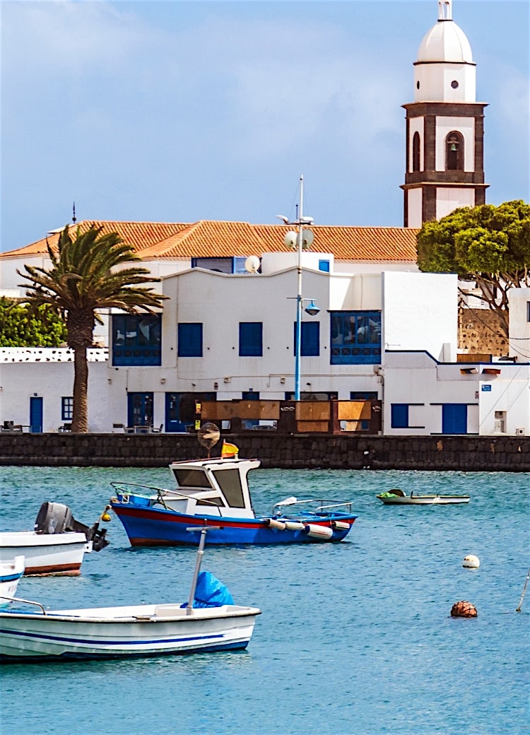Traditional fishing boats in Lanzarote harbour with waterfront buildings, palm trees and calm turquoise waters.